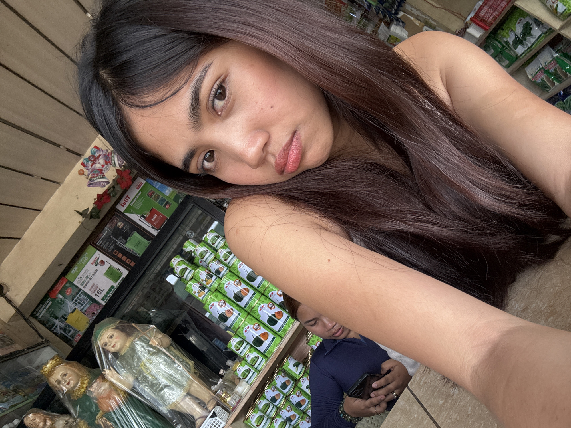 A young woman with long, dark hair and light makeup takes a selfie. She is in the foreground, with her head and shoulders filling much of the frame. Behind her, stacked cans of what appear to be food items are visible on shelves. To the left of the shelves, two religious statues are displayed, one appearing to be a woman in a green dress with a gold crown, and another smaller statue of a child. Further to the left, a man in a dark blue shirt is sleeping, holding a smartphone in his hand. The scene appears to be inside a small shop or market stall. This scene is located in Tagum City, Philippines.