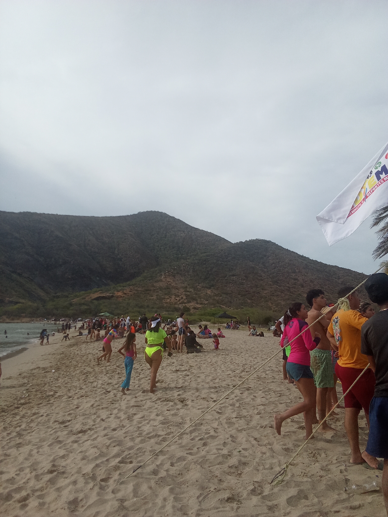 The image depicts a crowded beach scene in Juan Griego, Venezuela. Several people are on the sandy shore, some swimming in the water, while others are standing or walking along the beach. The background features a large, mountainous landscape under an overcast sky. A white flag with text and graphics is positioned to the right of the frame, near a group of people, and a rope is stretched across the foreground.