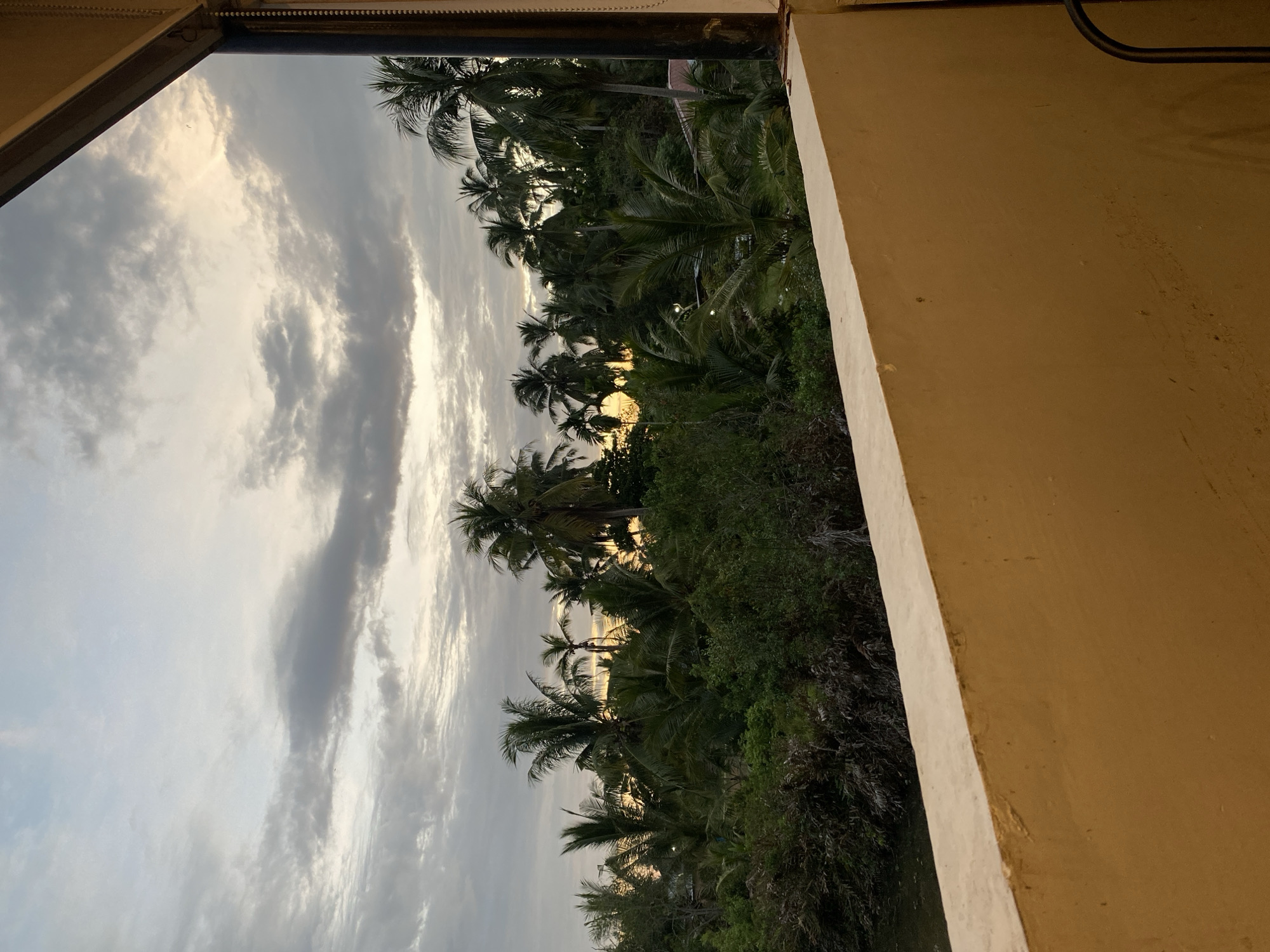 The image depicts a view from a building in Tanjongkapai, Indonesia. Through a window or opening, a tropical landscape is visible. Palm trees dominate the scene, silhouetted against a sky filled with clouds. The light suggests either sunrise or sunset. The building's structure, with its beige-colored wall, frames the external view.