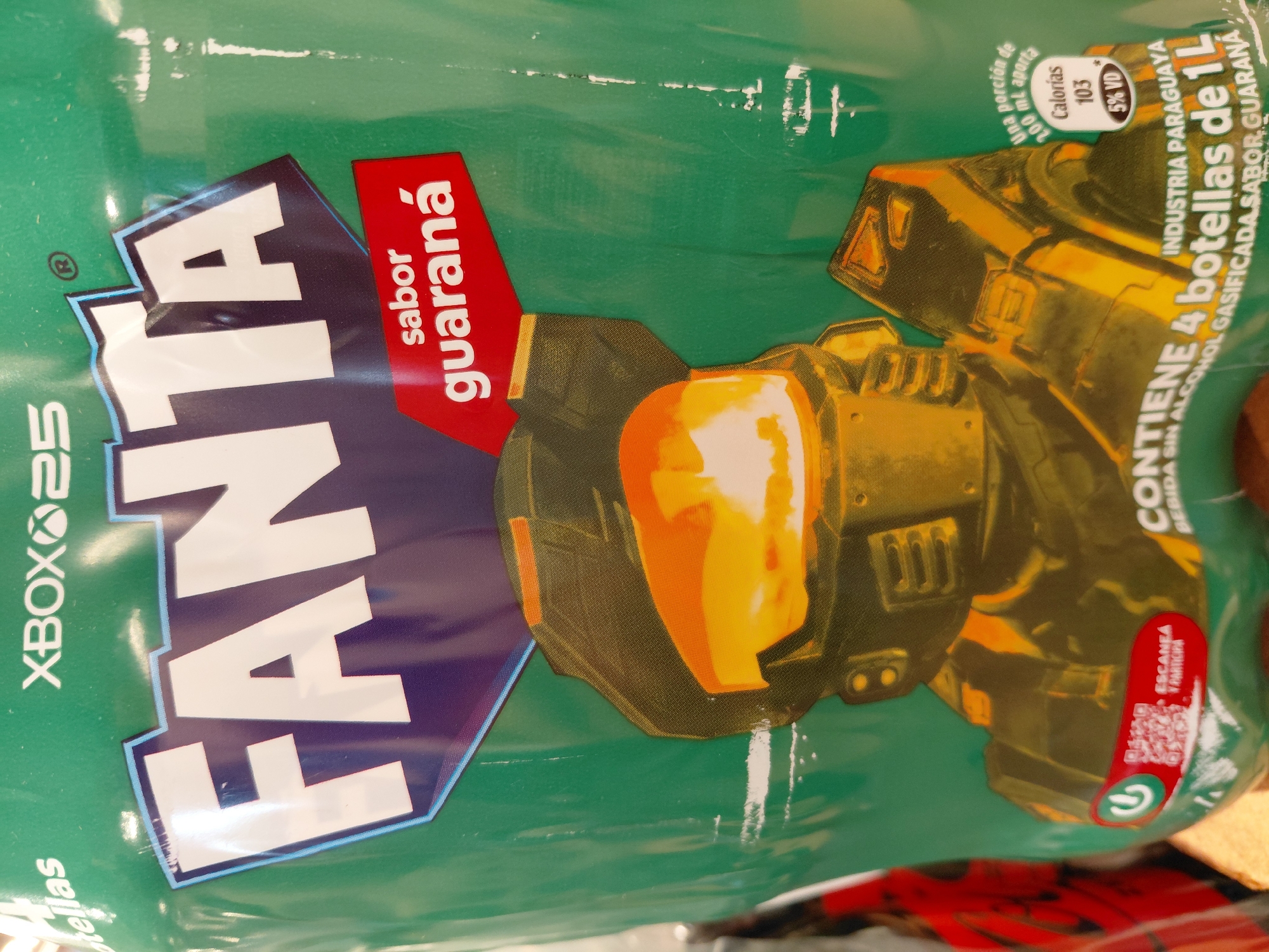 This media file is a close-up photograph of a green plastic bottle or packaging for a Fanta Guarana drink. The packaging features a prominent graphic of a character in golden armor, reminiscent of the Master Chief from the Halo video game franchise, with an orange visor. The Fanta logo is displayed in large, white, stylized letters with a blue outline. The words "sabor guaraná" are written vertically in white text within a red diamond shape.
Additional text on the packaging includes "XBOX 25," suggesting a collaboration with Xbox. Nutritional information is visible, stating "Una porción de 200mL aporta Calorías 103" and "5% VD" (Valor Diario/Daily Value). Another section reads "INDUSTRIA PARAGUAYA" and "CONTIENE 4 botellas de 1L BEBIDA SIN ALCOHOL GASIFICADA SABOR GUARANA," indicating it's a 4-liter pack from Paraguay, a carbonated, non-alcoholic guarana-flavored drink. A red oval contains a QR code and the text "ESCANEA Y PARTICIPA" (Scan and Participate). The overall impression is a promotional item, likely from a retail environment. The lighting suggests indoor conditions, and there are some minor scuff marks on the plastic surface.