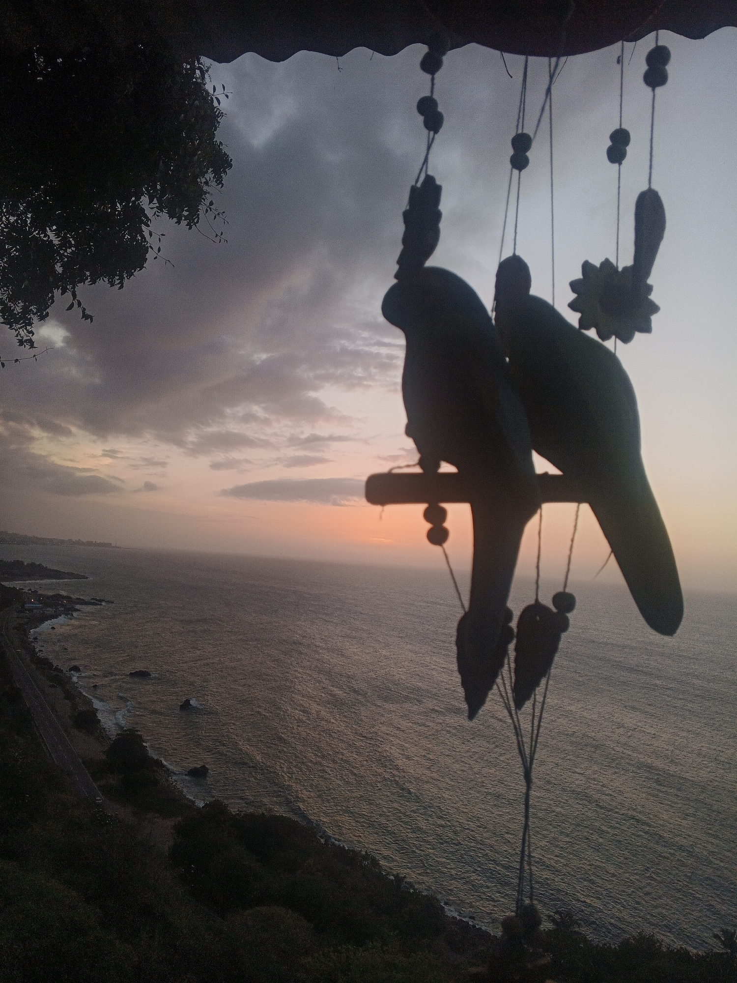 A silhouette of a wind chime with two bird figures and a flower ornament hangs in the foreground against a twilight sky and a calm ocean. The wind chime is suspended from what appears to be a roof or overhang. The sky displays a gradient of colors, transitioning from a pale yellow or orange near the horizon to a muted purple and gray towards the top, with scattered clouds. The ocean stretches out towards the horizon, its surface rippled by a gentle breeze. Along the coastline, a road is visible, winding its way through hilly terrain covered with vegetation. The overall lighting suggests it is either sunrise or sunset. The location is listed as Unknown, Unknown.