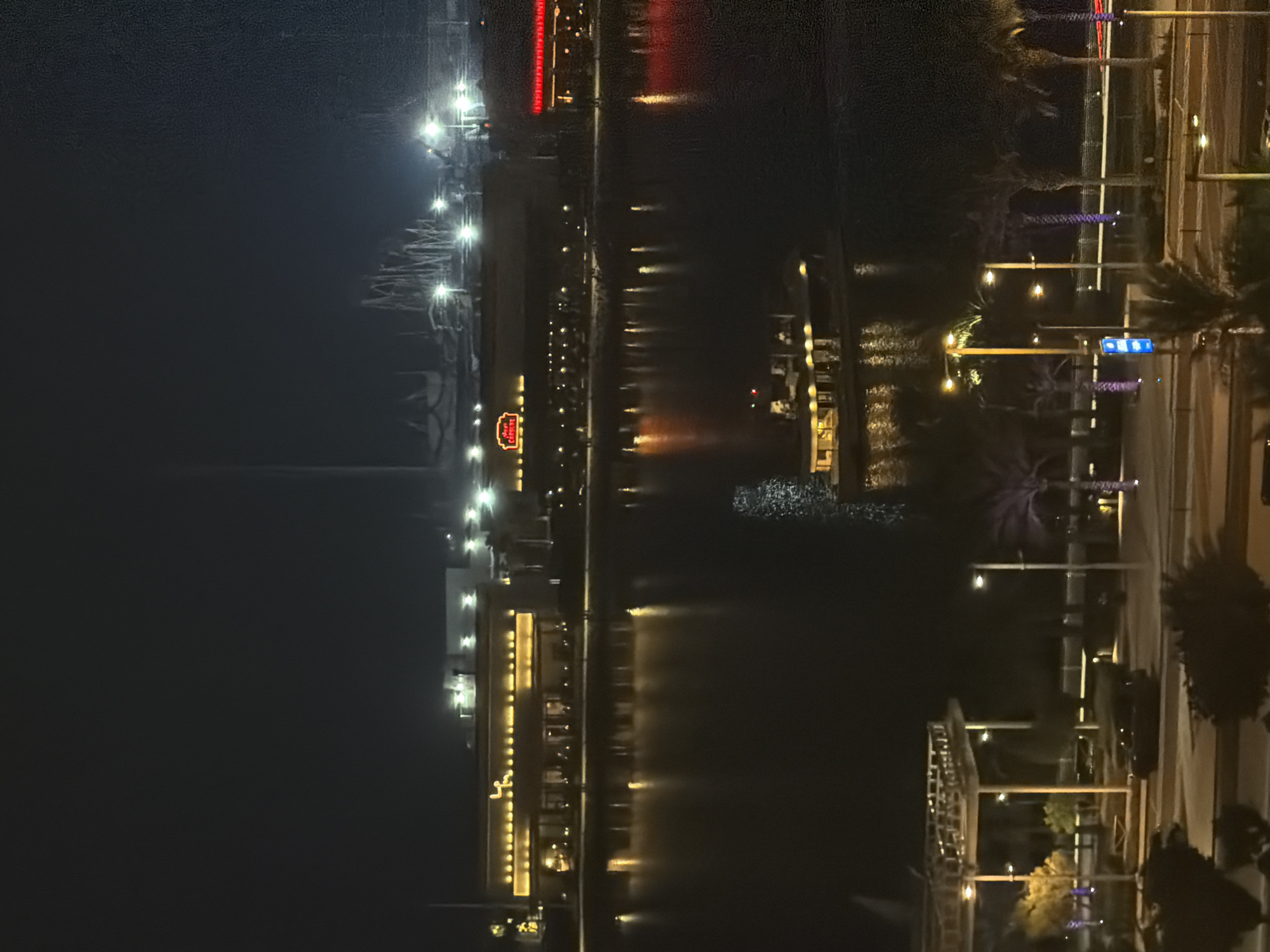 An aerial night-time view of a waterfront area in an unknown city. In the foreground, a dark road runs horizontally with several streetlights and palm trees lining the median. A black car is parked on the right side of the road. Beyond the road, there's a paved area with more palm trees and a covered structure. The middle ground features a calm body of water with reflections of lights from buildings on the opposite bank. A boat with illuminated windows is visible in the water. Across the water, a row of buildings is illuminated by warm yellow and red lights. One building has a sign that reads "THE CHEESECAKE FACTORY". In the background, silhouetted roller coasters and structures are visible against a dark sky, with several bright lights illuminating the amusement park. The overall scene is urban, with a focus on illuminated architecture and entertainment venues.
