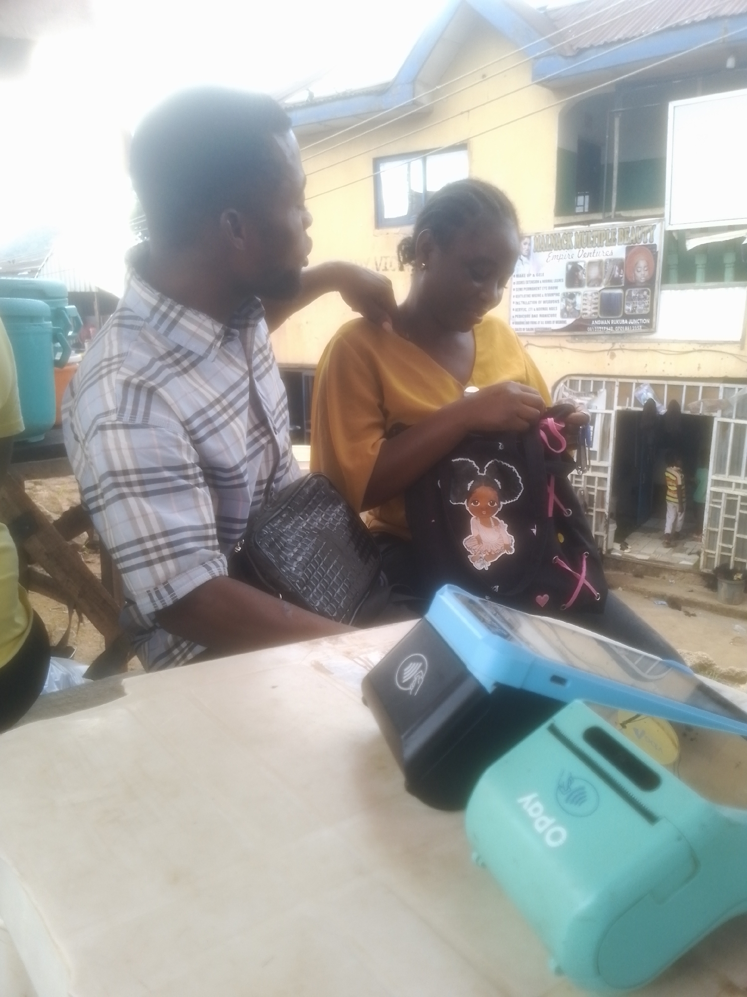 In an outdoor, urban setting, a Black man wearing a plaid shirt is seen from the chest up, leaning his left arm on the shoulder of a Black woman. The woman, facing to her left and looking down, wears a yellow top and is holding a black bag with pink laces and a cartoon image of a young girl. In the foreground, two payment terminals, one black and one teal with "OPay" branding, are on a light-colored surface, possibly a table. Behind the people, a building with a yellow facade and a sign for "MAINACK MULTIPLE BEAUTY Empire Ventures" is visible. The environment appears to be a street with some greenery and a white fence in the background.