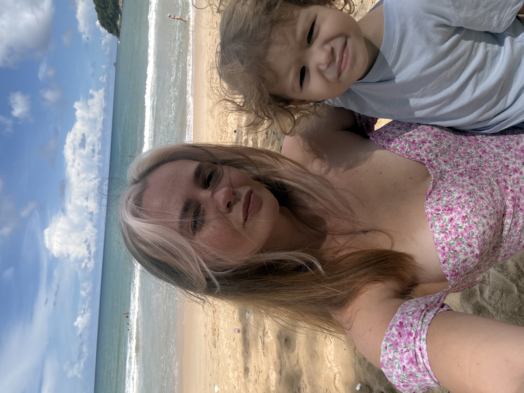 A woman with blonde and brown hair, wearing a pink floral off-the-shoulder top, takes a selfie on a beach with a young child. The child, wearing a light blue t-shirt, is held close to the woman. The beach is sandy with some litter visible. In the background, the turquoise ocean meets the horizon under a partly cloudy blue sky. A few people are in the water, and a green, tree-covered hill is visible in the distance on the right side of the frame. The image appears to have been taken in Choeng Thale, Thailand, given the tropical beach scenery.