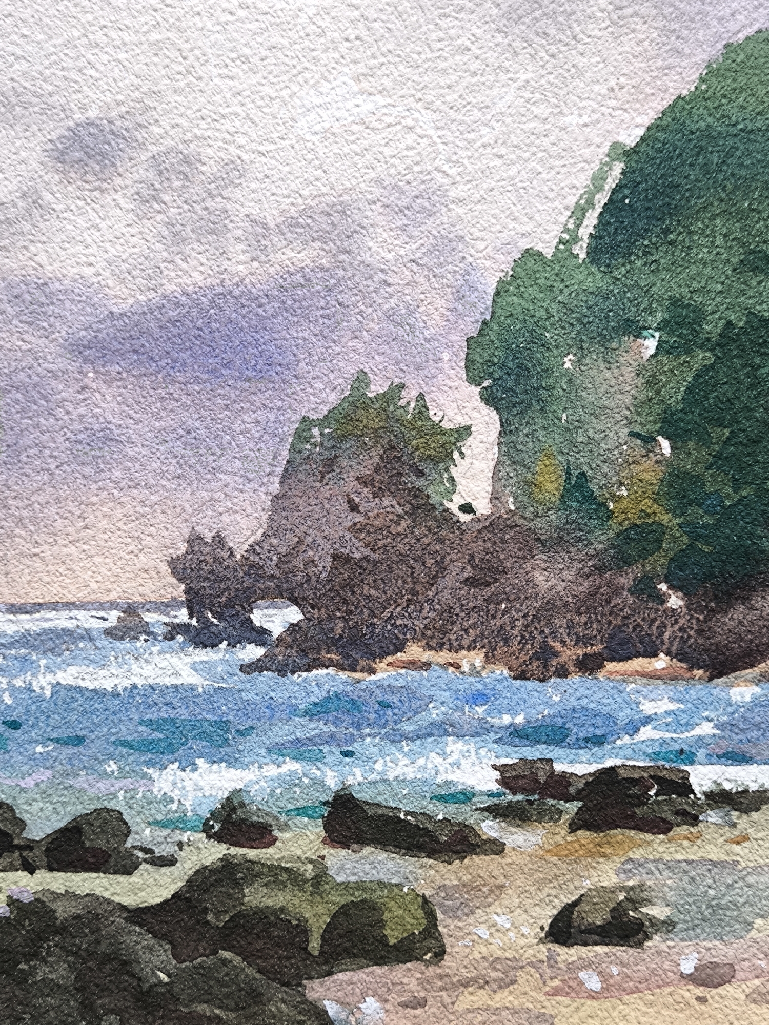 This is a watercolor painting depicting an unknown coastal location. The upper portion of the scene shows an overcast sky, rendered in light grey and pale lavender hues. To the right, a large, steep cliff face covered in dark green vegetation extends downwards towards the sea. In the midground, a series of dark brown and grey rocky formations project into the water, with one prominent rock structure displaying a distinct natural archway at its base. The ocean, depicted in shades of blue and turquoise, features white-capped waves actively breaking against the rocks and along the shoreline. The foreground reveals a rocky beach area composed of sand in varying tones of beige and brown, interspersed with scattered dark boulders and smaller rocks partially submerged in the shallow water. No individuals or man-made structures are visible.