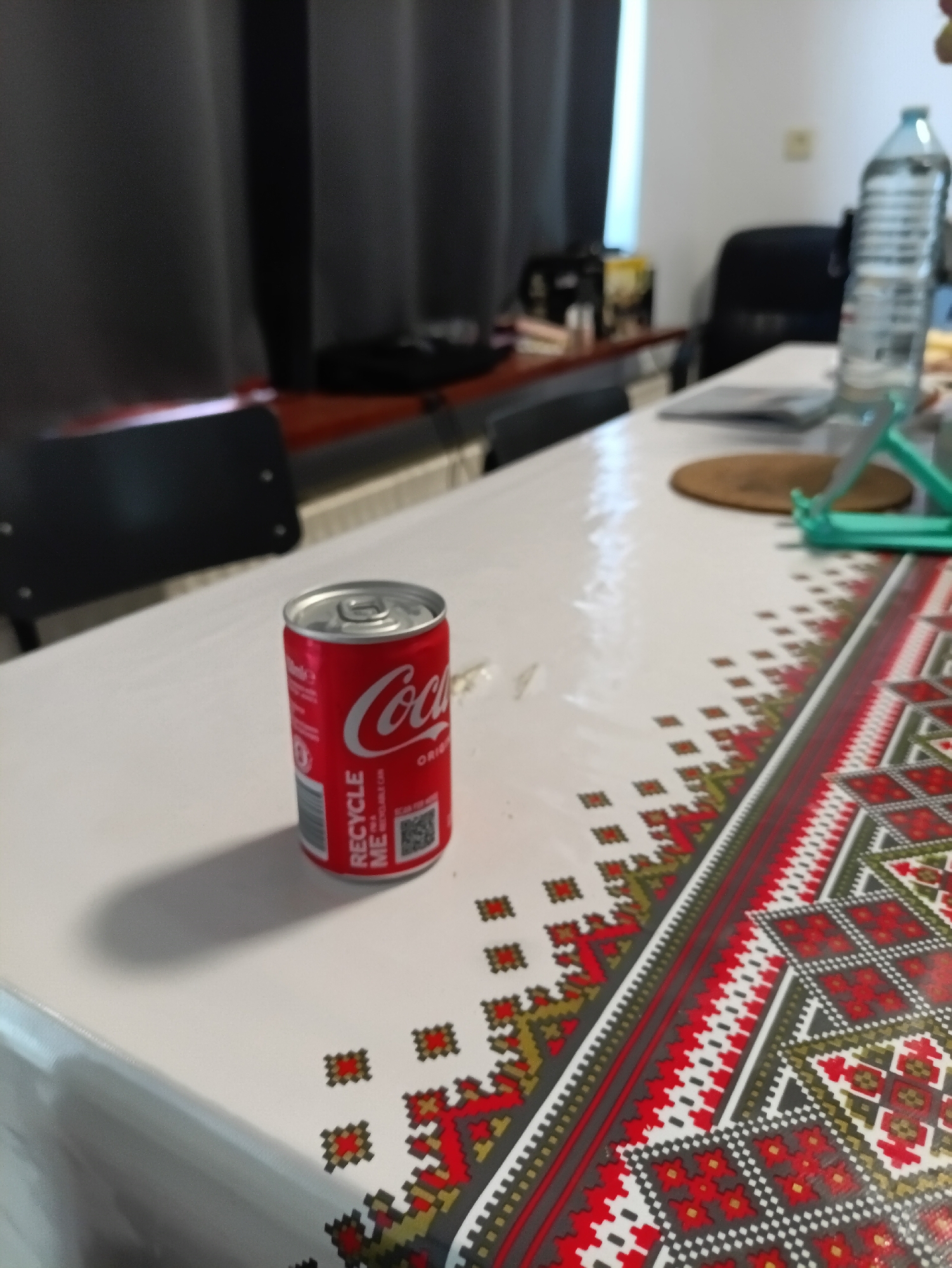 A Coca-Cola can is placed on a white, patterned tablecloth. The can is red with the Coca-Cola logo in white. The tablecloth has a border with red, white, and black geometric patterns. In the background, a water bottle stands on the table, and a chair and curtains are visible. The scene appears to be indoors, possibly in a home or office setting. There is no information available to determine the location in the city of Zwartewaal, Netherlands.