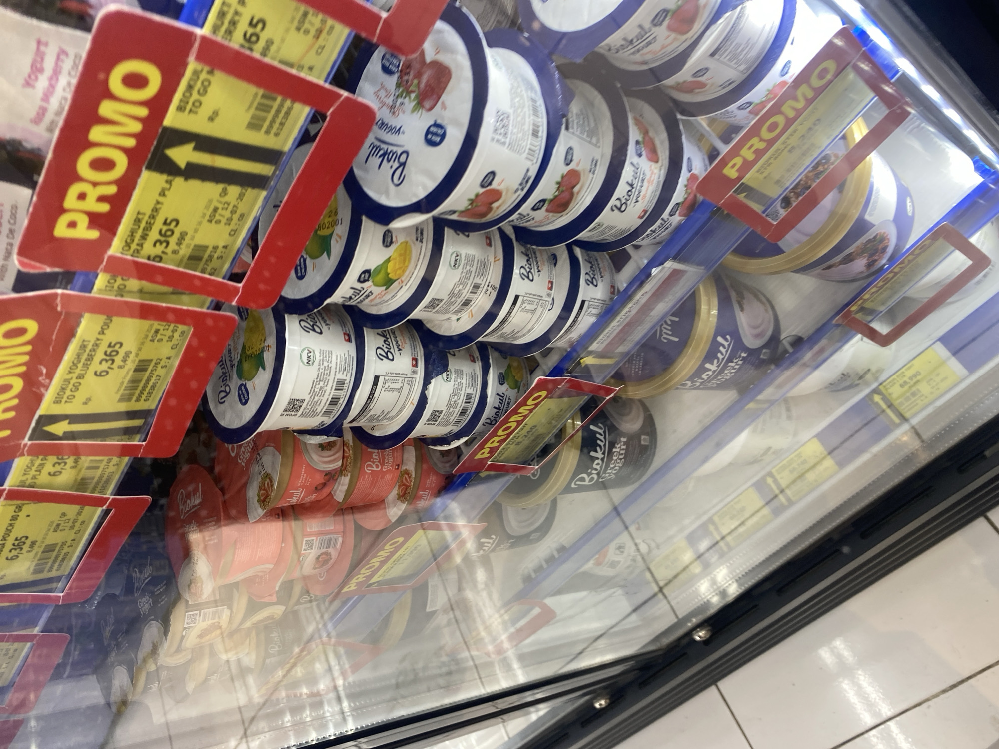 The image shows a refrigerated display case in a supermarket, likely in Watampone, Indonesia, filled with various yogurt products. Prominently displayed are red and yellow "PROMO" signs indicating special offers on the yogurts. The products appear to be individual serving cups of yogurt, with brands like "Biokul" visible. Some cups feature images of fruits like strawberries and mangoes, suggesting different flavors. The lighting suggests an indoor environment, typical of a retail store. There are no people visible in the image. The overall scene depicts a typical retail environment showcasing a selection of dairy products on promotion.