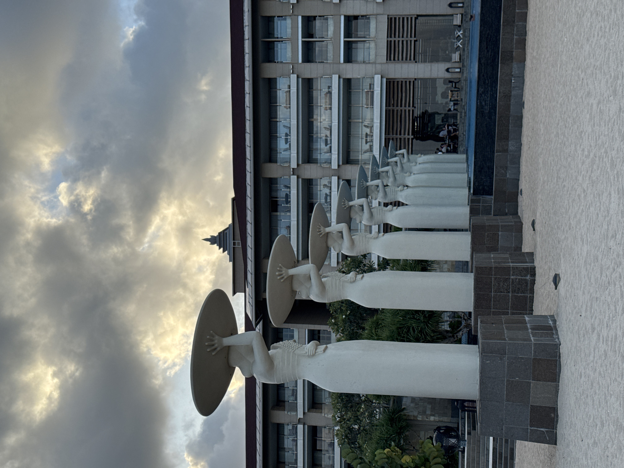 The image shows a row of white sculptures depicting female figures holding circular objects above their heads, likely umbrellas or parasols. The sculptures are located on a sandy area, presumably a beach, with a modern building in the background. The building has a multi-story facade with multiple windows. Lush greenery surrounds the base of the sculptures and the building. The sky is overcast with dramatic cloud formations. The location is Kuta Selatan, Indonesia, as the sculptures are known to be located there. A swimming pool is seen behind the sculptures. People are visible on the building's deck, behind the sculptures and the pool.