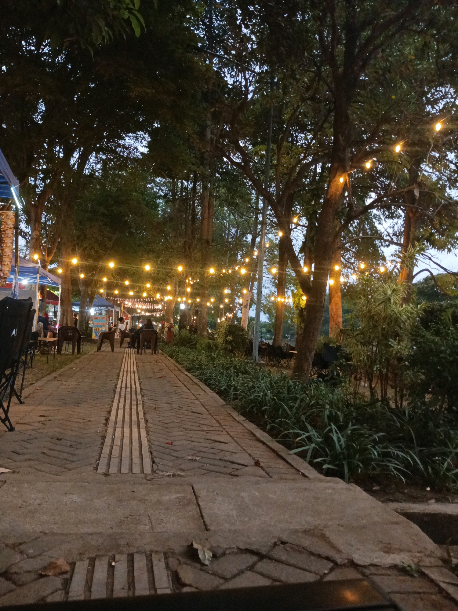The image depicts a pathway lined with trees, illuminated by strings of warm, yellow fairy lights. The pathway is paved with stone, with a distinct section of raised, ridged tactile paving. On either side of the pathway, there are areas with tables and chairs, suggesting an outdoor dining or seating area. Several people are visible, seated at tables or walking along the path. Some are silhouetted against the lights, while others are more discernible. In the background, there are various structures that appear to be stalls or tents, and more trees creating a canopy overhead. The lighting suggests it is dusk or evening. This scene is likely from the area near Alun-Alun Tulungagung in Tulungagung, Indonesia, a common location for evening gatherings and food stalls, especially when decorated with lights.