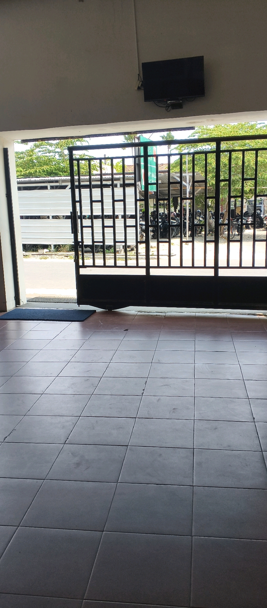 The image depicts an outdoor scene, possibly a covered walkway or entrance, leading to an open gate. The foreground is a tiled floor in shades of grey and brown. A dark blue mat is visible near the opening. Beyond the mat, an open black metal gate with a grid pattern allows a view of the outside.
The outside area features a street with parked motorcycles, suggesting a parking area or a street-side location. A large white truck with a ribbed side is parked on the left. Trees with green foliage and some buildings with tiled roofs are visible in the background, indicating an urban or semi-urban setting. A black television is mounted on a grey wall above the gate.
The location is identified as Planggu, Indonesia. The image is taken from a low angle, looking outwards through the opening. There are no people visible in the image. The overall impression is that of an entrance to a property or business in Planggu.