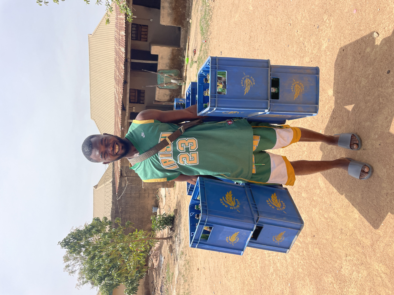 A man stands outdoors on a sunny day in Barakin Akawo, Nigeria, smiling broadly directly at the camera. He is carrying a significant load of blue plastic crates, with two stacked on each side of his body, containing bottles.
The man has dark skin and a short beard. He is wearing a green basketball-style jersey with yellow trim, featuring the number "23" in white and a "NIKE" logo on the left shoulder. He pairs this with yellow and white shorts and gray slides. A dark shoulder bag, with a pattern that includes "LOUIS VUITTON" text, is slung across his chest.
The blue crates are clearly branded with a golden crest logo, resembling wheat or barley, and the words "PREMIUM BREW" and "LITE" are visible on their sides, indicating they hold bottled beverages.
The scene is an unpaved, reddish-brown dirt area. In the background, there is a low-rise building with a corrugated metal roof, suggesting a residential or commercial structure. A green wheelbarrow is visible near the building. Sparse vegetation, including a small tree or bush, dots the dusty landscape. The bright, clear sky suggests it is daytime. The man appears cheerful despite his heavy load.