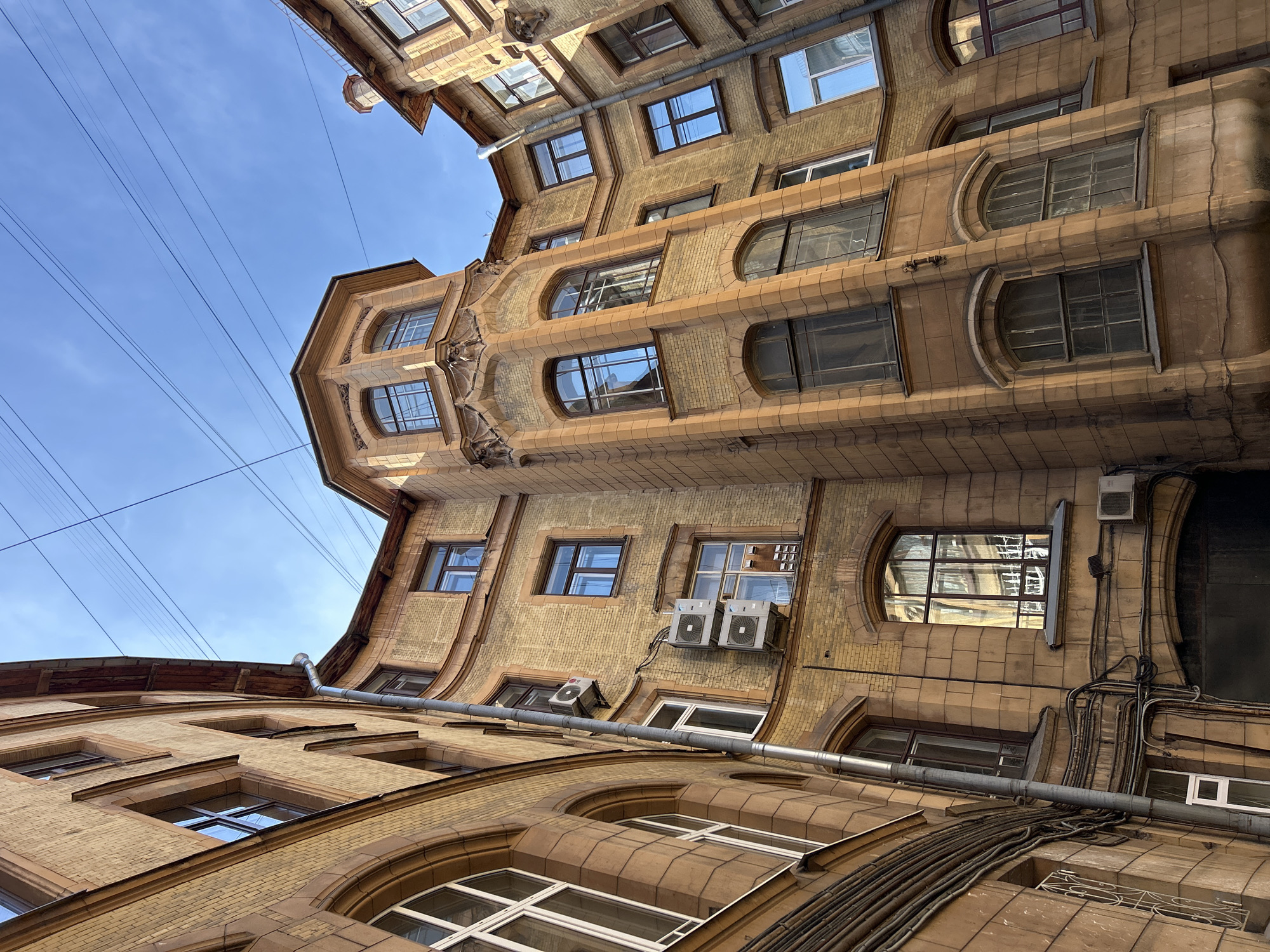 An outdoor, low-angle, full shot shows the facade of old, yellow brick buildings with brown trim surrounding a narrow courtyard. The sky above is clear and blue, crisscrossed by multiple black electrical wires. The buildings have numerous windows, some arched and some rectangular, with dark frames. Several air conditioning units are attached to the exterior walls. A large, dark metal gate is visible at the bottom center. The architecture suggests an older European city, possibly St. Petersburg.
