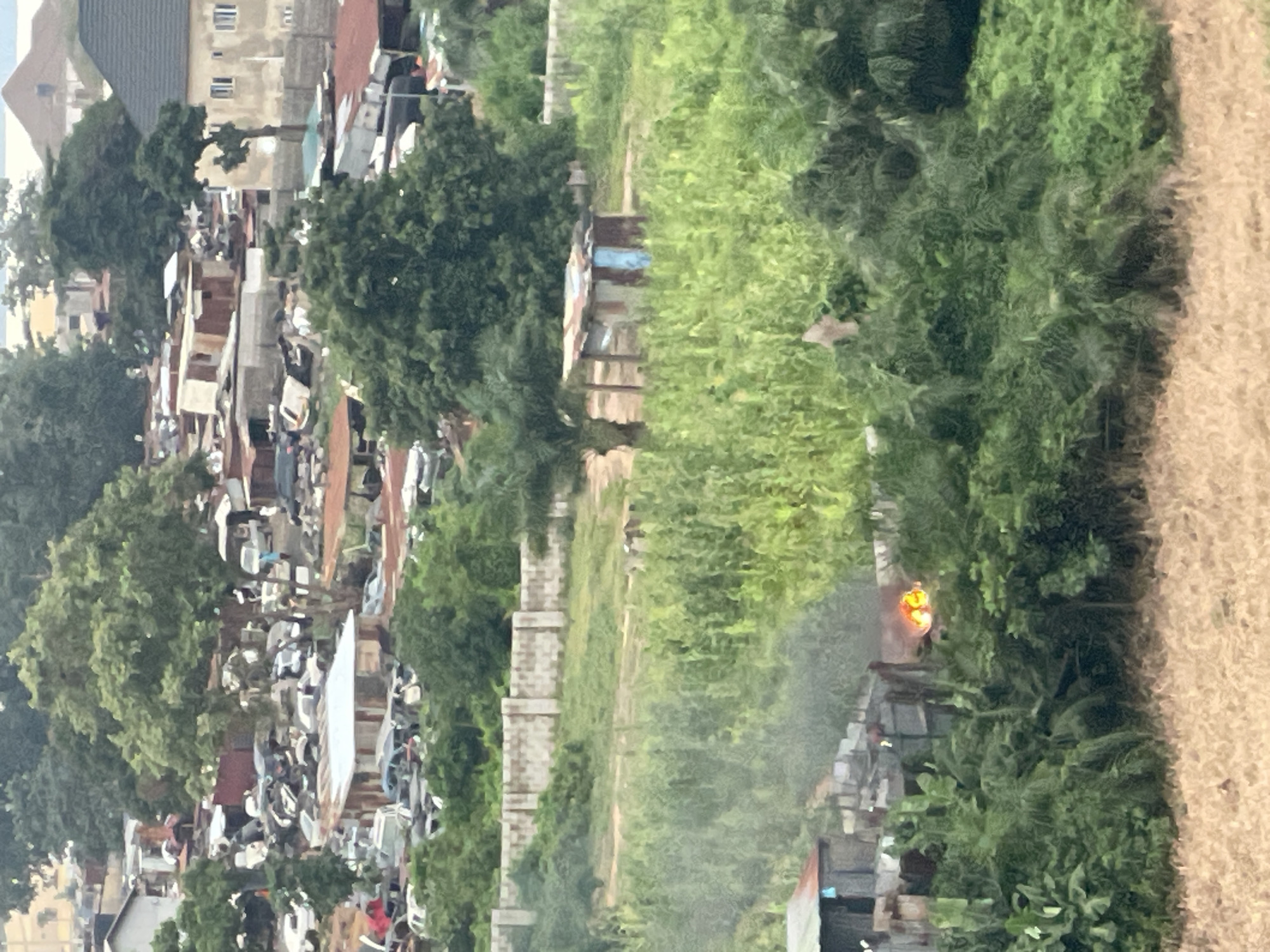 The image depicts an outdoor scene within a city that appears to be Abuja, Nigeria, characterized by a mix of residential and industrial elements. In the foreground, dense green foliage, possibly a field or overgrowth, occupies the lower portion. Towards the middle ground, a small, makeshift structure, likely a shack or informal dwelling constructed from corrugated metal and wood, is visible. Adjacent to this structure, a fire is actively burning, emitting smoke and orange flames, suggesting some form of controlled burning or an uncontrolled incident. Beyond this, a concrete block wall stretches horizontally, separating different areas of the landscape. In the background, a densely packed area of buildings and structures is visible, including what appears to be a scrapyard or a car park with numerous vehicles, some of which are partially dismantled or in a state of disrepair. The overall impression is one of an urban fringe environment where informal settlements and industrial activities coexist with residential housing.
