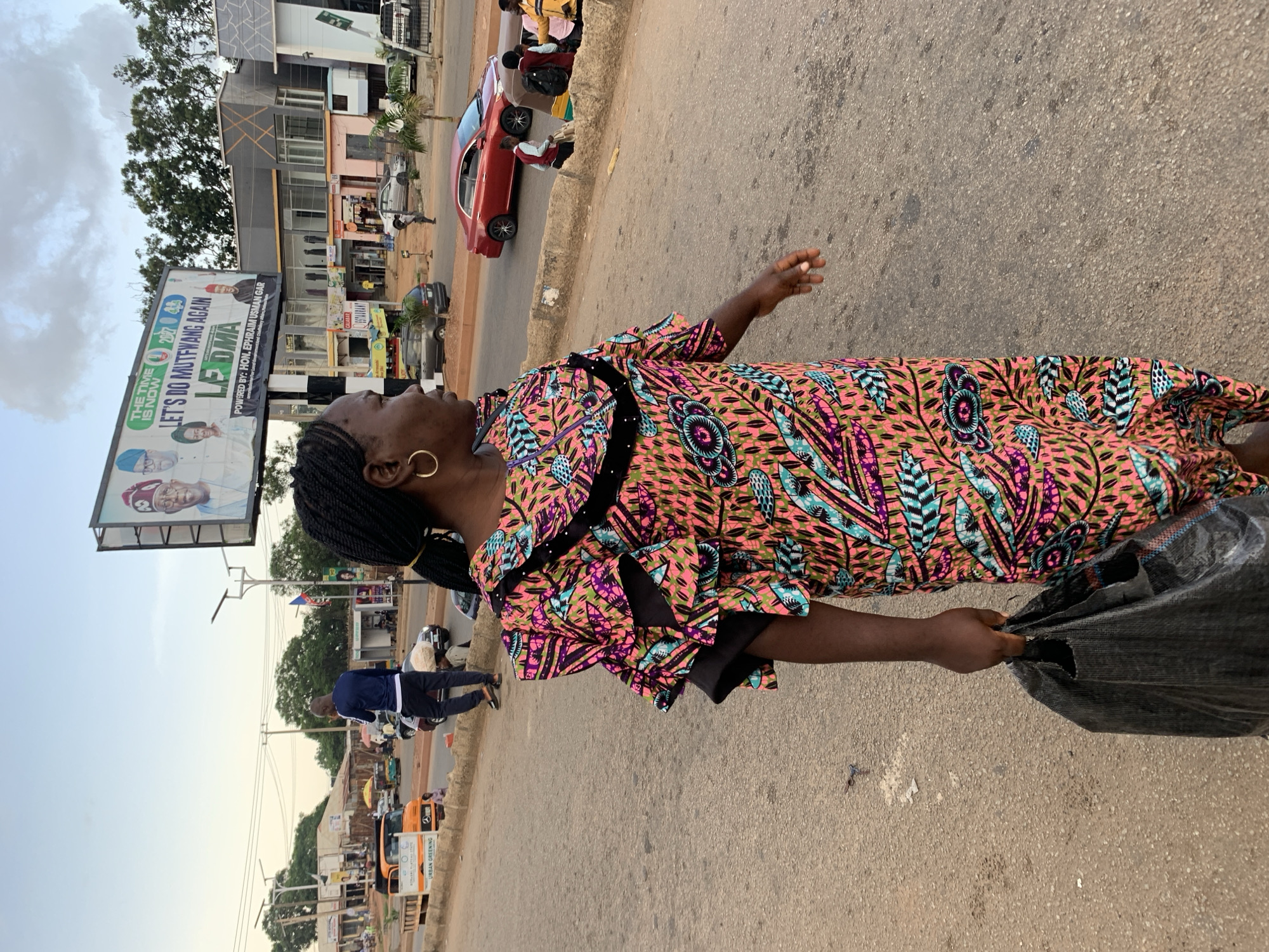 In this outdoor, vertical shot, a Black woman is captured from the chest up, standing on a paved street with a billboard behind her. She wears a vibrant, patterned dress in shades of pink, blue, and purple, with ruffle details on the sleeves and neckline. A large, gold hoop earring adorns her left ear. Her right arm is extended, holding a crumpled black plastic bag. Her gaze is directed upwards and to her left, with a slight smile on her face, suggesting a moment of observation or anticipation.
The background reveals a busy street scene in Jos, Nigeria. A billboard advertising "THE TIME IS NOW" and "LET'S DO MUTFWANG AGAIN" with images of politicians dominates the left side of the frame. Further down the street, various shops and businesses are visible, including one with the word "CALABAR" on its signage. Several vehicles, including cars and motorcycles, are present on the road, indicating a typical daytime activity. The weather appears to be overcast with a pale sky. The overall impression is that of a person going about their day in a bustling urban environment.