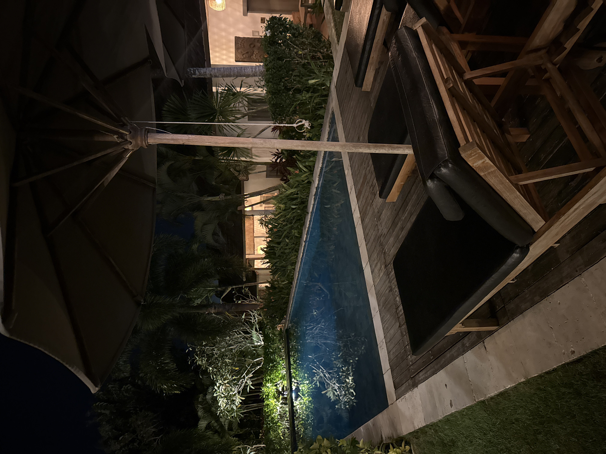 The scene captures an outdoor setting, likely in a private garden or courtyard, at night in Ungasan, Indonesia. A swimming pool runs lengthwise through the frame, reflecting light and the surrounding foliage. On the right of the pool, wooden decking supports lounge chairs with black cushions. To the left, the edge of an open umbrella dominates the upper left corner of the frame. Various plants, including palm trees, flank the pool. A structure is visible in the background, featuring architectural details and illuminated by warm-toned lights. The lighting creates a contrast between the brighter building and the darker foliage.