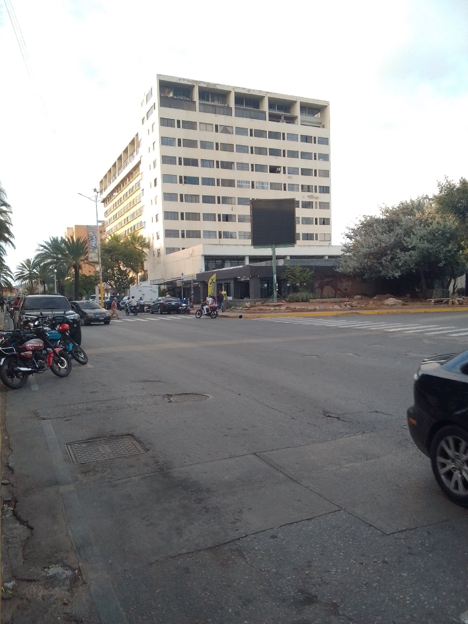 The image shows a street scene in the city of La Tagua, Venezuela. A large, light-colored, multi-story building dominates the background. In the foreground, several motorcycles are parked along the side of the road. A black SUV is also present. The road is paved, and there are crosswalk markings indicating an intersection. Trees and palm trees are visible, suggesting a tropical or subtropical climate. The sky is bright, indicating daytime. The scene appears to be a relatively busy urban environment with traffic and parked vehicles.