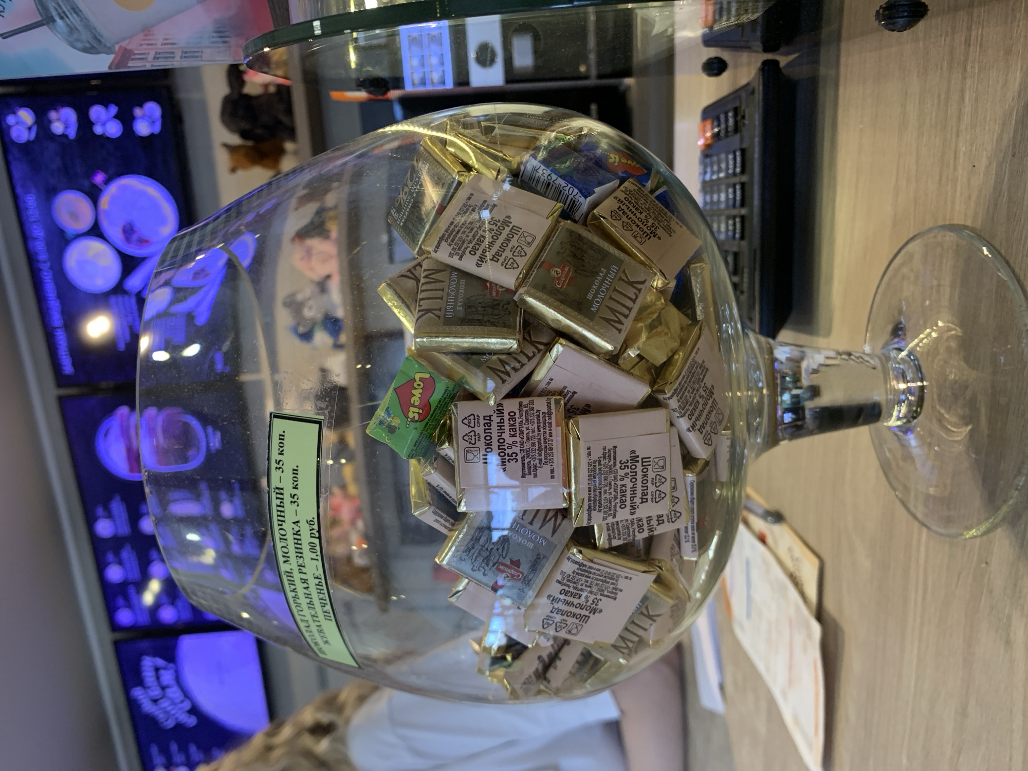In a dimly lit establishment, possibly a cafe or shop, a large glass bowl overflows with individually wrapped chocolates. The bowl sits on a wooden counter, where a calculator is also visible. In the background, screens display illuminated graphics, suggesting a modern retail environment. A portion of a person's arm and shoulder, clad in a white shirt, is visible to the left, indicating a customer or staff member. The overall scene is an indoor shot, capturing a retail display of sweets. This image was captured in Sluck, Belarus.