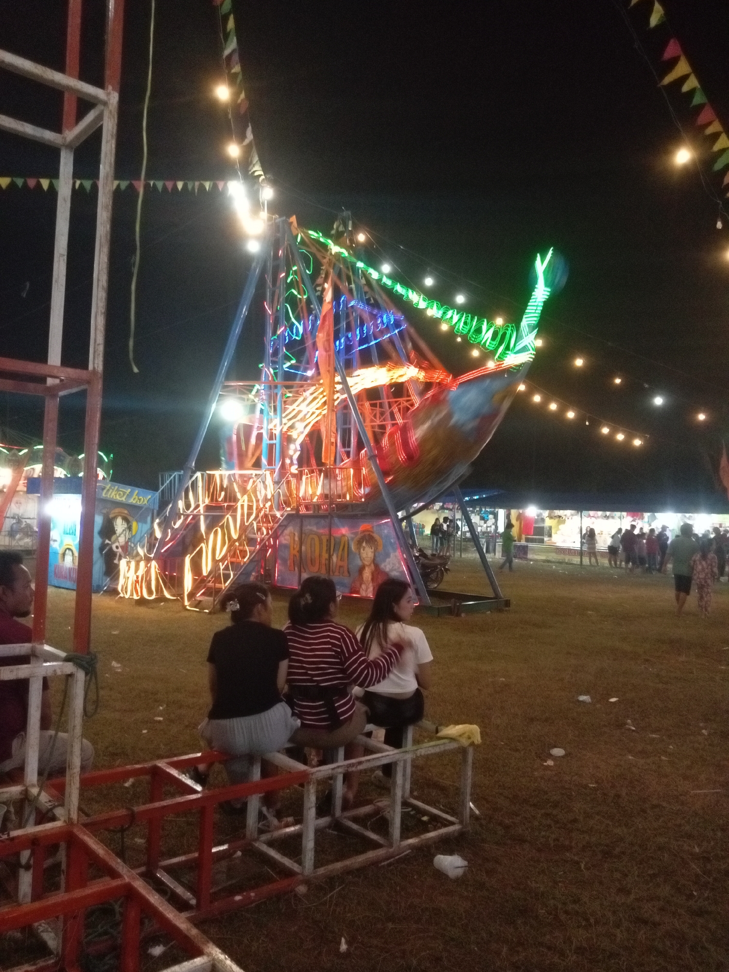 An outdoor amusement park attraction, likely a spinning boat ride, is illuminated with vibrant neon lights at night. The ride features a large, boat-shaped structure with a colorful, multi-colored light display. In the foreground, several individuals are seated on a metal structure, facing away from the camera and towards the ride. The ground is covered in dry grass, with scattered litter visible. In the background, various stalls and a crowd of people suggest a bustling event. This scene is characteristic of a temporary fair or festival, possibly located in the city of Batam, Indonesia, given the context of such attractions in the region.
