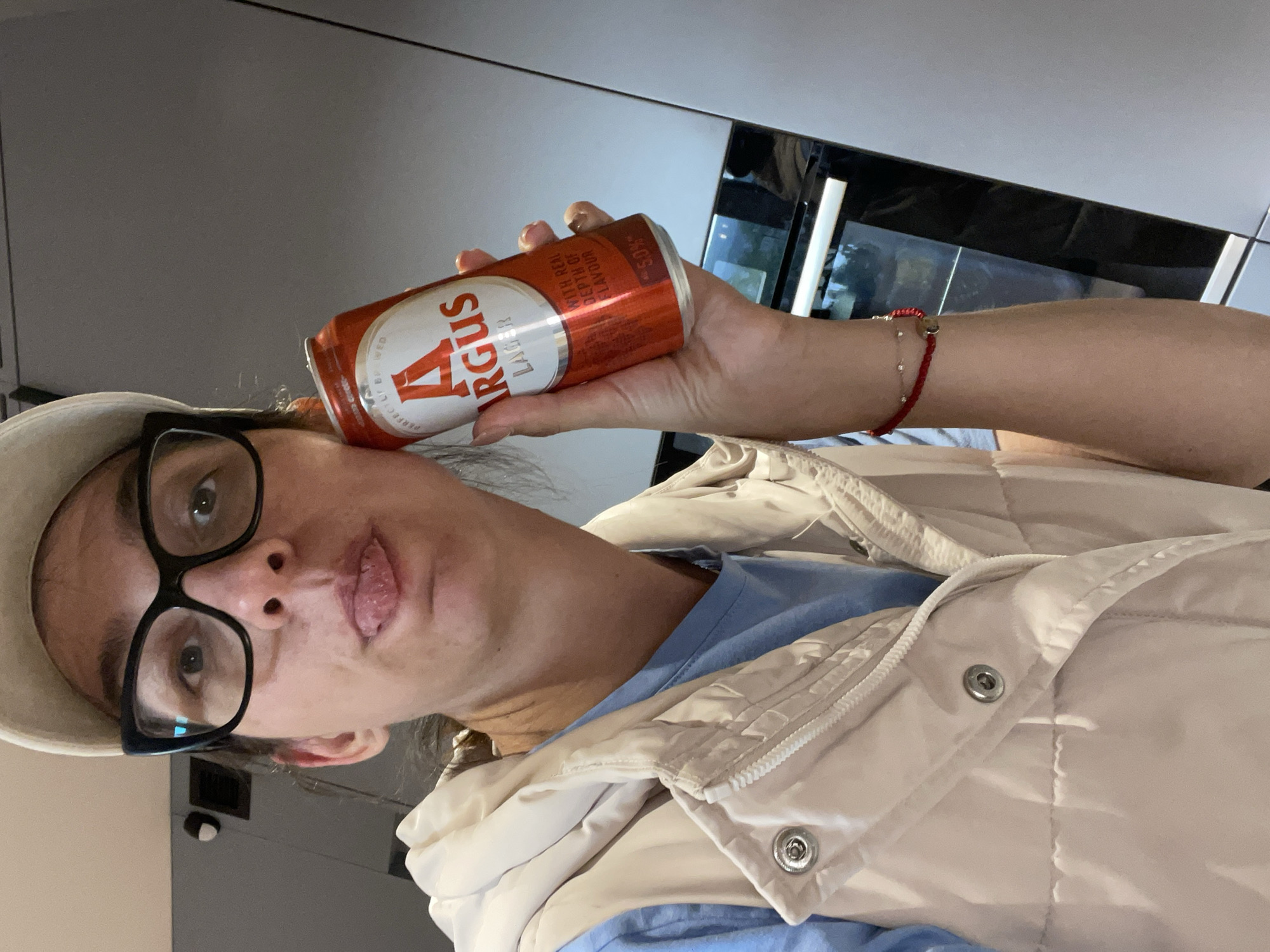 A woman is holding a can of Argus beer, a brand available at Lidl, next to her head as if it were a phone. She is wearing black-framed glasses, a beige hat, a blue shirt, and a light-colored padded vest. She is indoors. The location appears to be a kitchen, judging by the countertops and possibly an oven/microwave unit visible in the background. This is likely a domestic interior, but without street-level details, it's not possible to pinpoint a precise location within Sofia, Bulgaria.