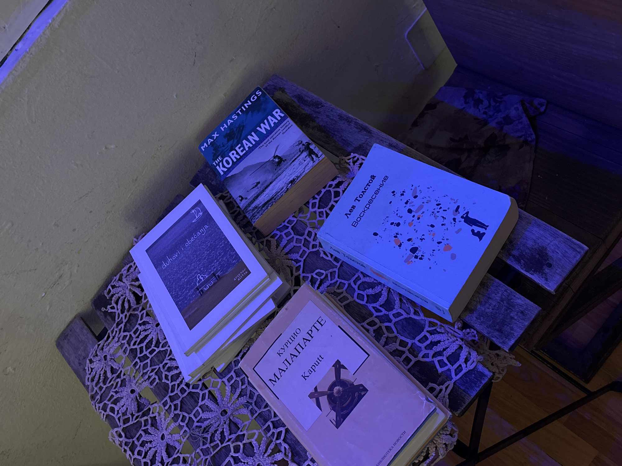 The image depicts four books placed on a small wooden table covered with a lace doily. The books, from left to right, are "duhovi i obećanja" by Arsenov Aleksa, "THE KOREAN WAR" by Max Hastings, "Воскресение" by Лев Толстой (Leo Tolstoy), and "Kaputt" by Curzio Malaparte. The table is situated in a room with a yellow wall visible in the top left corner and a dark corner in the top right and bottom right, suggesting a partially illuminated space.
The image was likely taken in Belgrade, Serbia because "duhovi i obećanja" and "Kaputt" by Curzio Malaparte are both published in Serbian language.