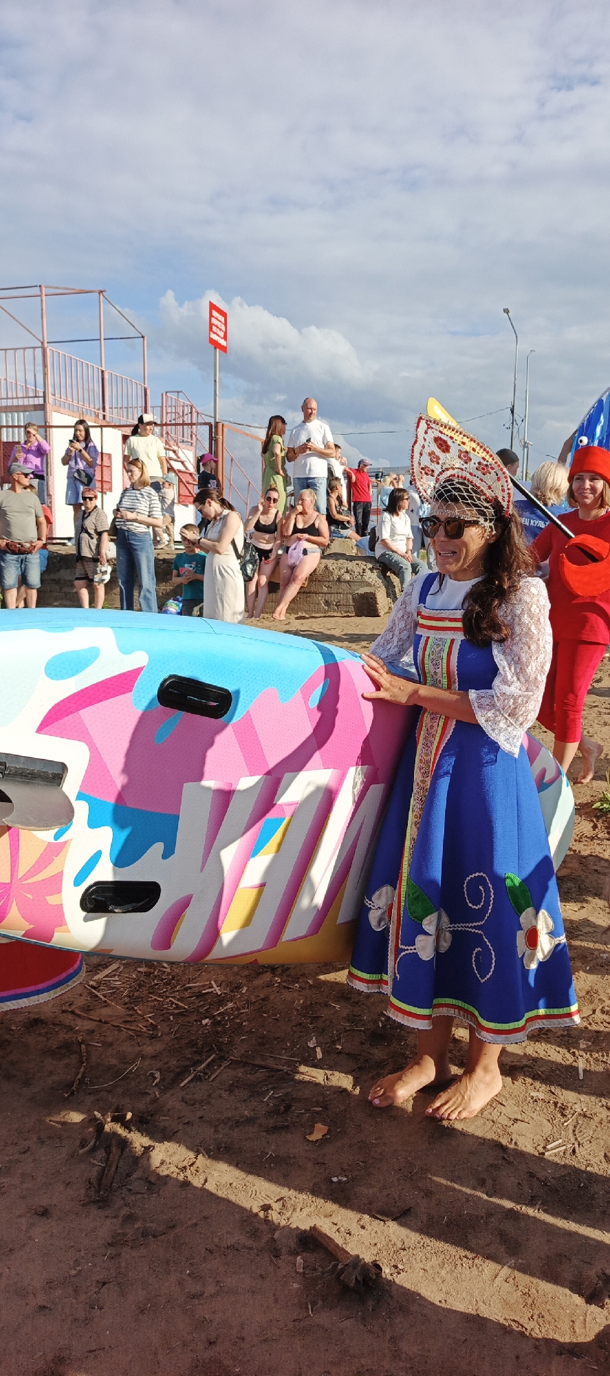 A woman in a blue traditional dress and ornate headwear stands next to a colorful inflatable object, possibly a watercraft, on a sandy surface. She is wearing sunglasses and appears to be smiling. Behind her, a crowd of people are gathered, some standing and some sitting on concrete structures. The background includes a light blue sky with scattered clouds, some railings and a red signpost. In the distance, there are buildings and a lamp post. The scene appears to be outdoors, possibly at a beach or recreational area in the city of Chaikovsky, Russia, given the presence of a traditional costume.