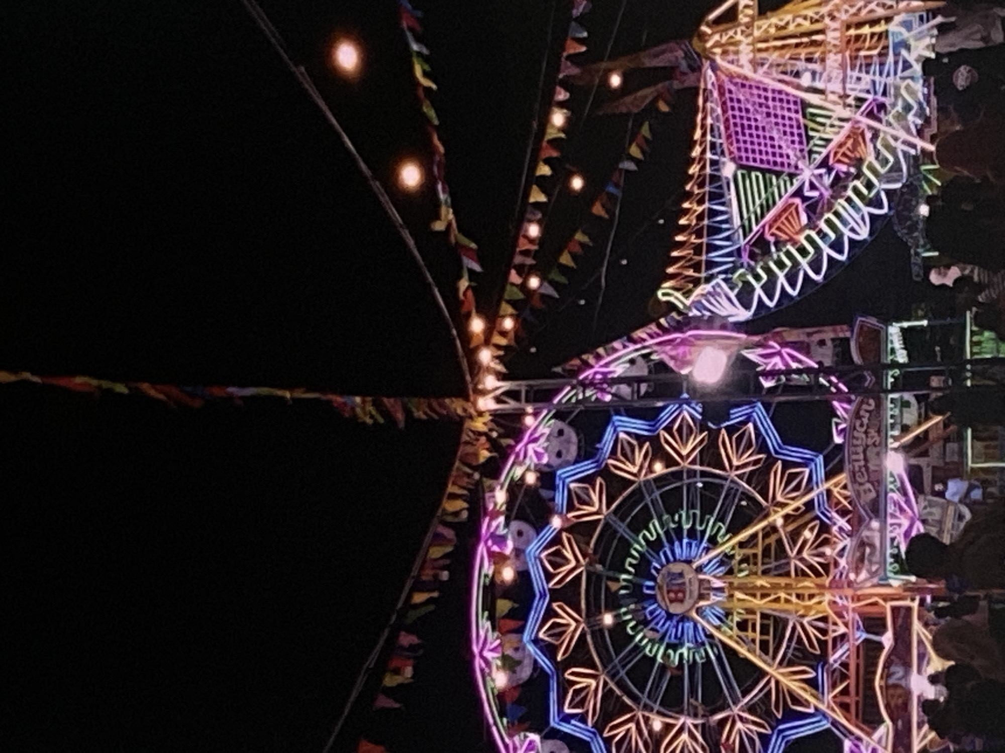 The image captures a nighttime scene at a vibrant carnival or fairground in Tebel, Indonesia. The foreground is populated by silhouettes of people, suggesting a lively gathering. Dominating the background are brightly lit amusement rides, specifically a large Ferris wheel adorned with neon lights in blue, pink, and yellow, and another ride with a curved structure also outlined in colorful neon. Numerous strings of small, colorful pennant flags are strung across the scene, along with incandescent bulbs that add to the festive atmosphere. The sky is dark, indicating it is nighttime. The overall impression is one of a bustling and illuminated night fair.