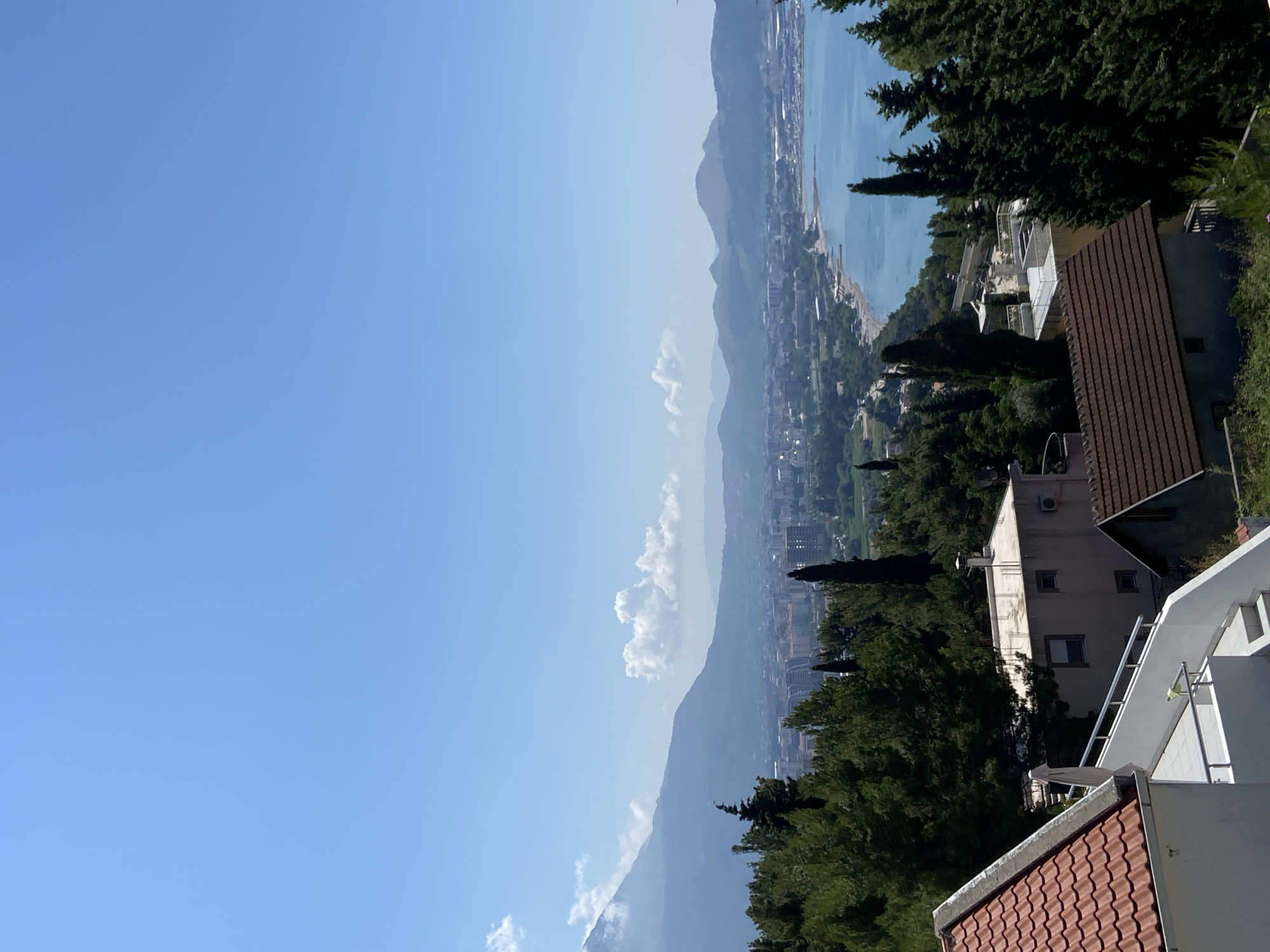 The image displays a panoramic view of the city of Šušanj, Montenegro, on a clear day with a vibrant blue sky. In the foreground, several rooftops of houses are visible, characterized by red and brown tiles. Beyond these houses, lush green trees and cypress trees dominate the landscape, interspersed with modern buildings and a few older structures.
In the mid-ground, the city of Šušanj unfolds, with a variety of buildings showcasing different architectural styles, including multi-story residential and commercial structures. A prominent feature is the coastline, where a tranquil blue sea meets a beach. The water appears calm and inviting, with a few scattered boats visible further out.
The background is dominated by a range of mountains that frame the city and the bay. The mountains appear hazy, suggesting a considerable distance. A few scattered white clouds are present in the sky, adding depth to the scene. The overall impression is one of a picturesque coastal town nestled between mountains and the sea, under a bright summer sky. There are no people or discernible actions taking place in the image.