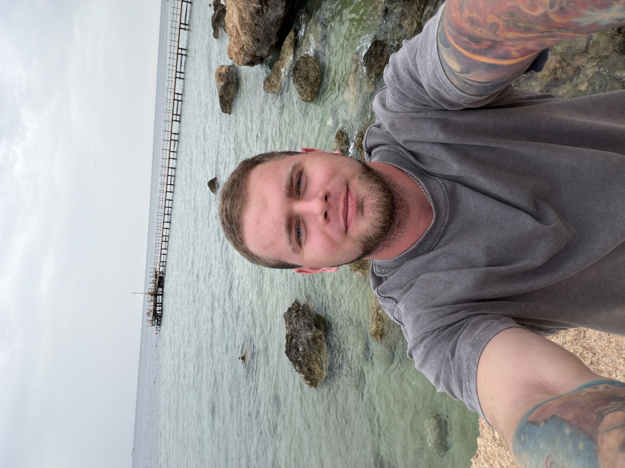 A man with a beard is taking a selfie near the beach. He is standing on a rock-covered shore, with a view of the ocean and a long pier extending into the water. The location is most likely in Sharm El Sheikh, Egypt, given the geographical features. The man is wearing a gray shirt and has tattoos on his arms. The sky is overcast.