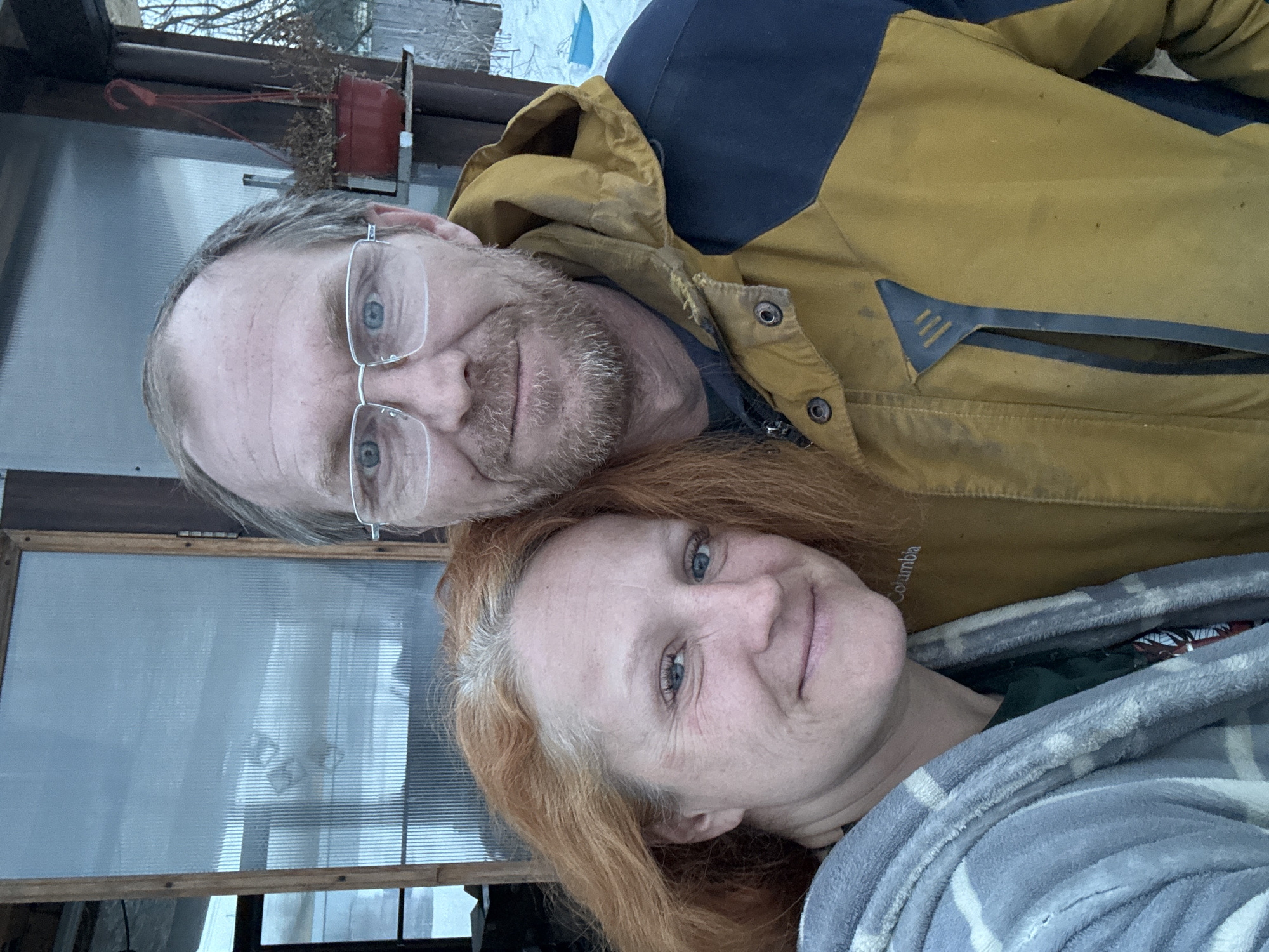 A selfie-style photograph captures a man and a woman outdoors in Chaikovsky, Russia. The woman, positioned on the left, has long reddish-blonde hair with visible gray roots, light blue eyes, and a slight smile. She is wearing a grey and white plaid patterned fuzzy robe over a dark green top. The man, positioned on the right and slightly behind the woman, has short light brown hair, a light brown beard, blue eyes, and wears silver-framed glasses, also displaying a slight smile. He is wearing a mustard yellow and navy blue two-tone winter jacket, which exhibits visible dirt marks. The immediate background features a wooden-framed structure with translucent ribbed plastic panels, possibly a greenhouse or shed. To the right of the man, a dark red plastic hanging pot containing withered plant material is situated on a shelf. The far background reveals bare tree branches against an overcast sky and patches of snow on the ground, indicative of a cold season. A blue plastic object, possibly a sled or container, is partially visible in the snow on the far right.
