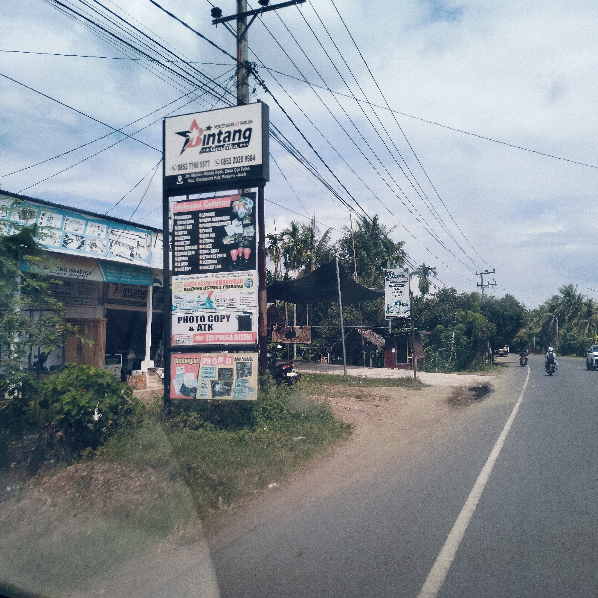 The image depicts a daytime street scene in Cottupah, Indonesia, showing a two-lane asphalt road with a white divider line, flanked by commercial establishments and lush tropical vegetation under a cloudy sky. Numerous overhead wires crisscross the sky, attached to a utility pole on the right side of the road.
On the left, a prominent multi-panel billboard advertises "Bintang Grafika," a printing and screen-printing service. The main panel displays the business name, phone numbers (0852 7766 5577, 0852 2020 9984), and address: "Jln. Medan - Banda Aceh, Desa Cot Tufah, Kec. Gandapura Kab. Bireuen - Aceh." Below this, a section lists extensive services under "Melayani Cetakan" (Serving Printing Services), including invitations, banners, business cards, pre-wedding photos, shop signs, digital flower boards, plaques, calendars, neon boxes, name tags, stamps, inscribed stones, stickers, T-shirt screen printing, mugs, keychains, brochures, passport photo printing, ID cards, certificates, photocopying, and more. Additional signs on the billboard announce an "Official Payment Counter for Electricity Bills & Prepaid," indicate "PHOTO COPY & ATK" (photocopy and stationery), and advertise "ISI PULSA DISINI" (top up credit here). To the left of the billboard is the "Bintang Grafika" shop building itself, with additional signage.
On the right side of the road, a shop covered by a dark canopy is visible, with dense greenery and palm trees behind it. Further down the road, another smaller sign, possibly for "Istana" (Palace), lists services like "Mobil" (car), "Ambal" (carpet), "Motor" (motorcycle), and "DLL" (etc.), suggesting a repair or general service shop. Several motorcycles are seen on the road, with at least two riders clearly visible heading away from the viewer. The road appears well-maintained, and the overall impression is one of a busy local commercial area.