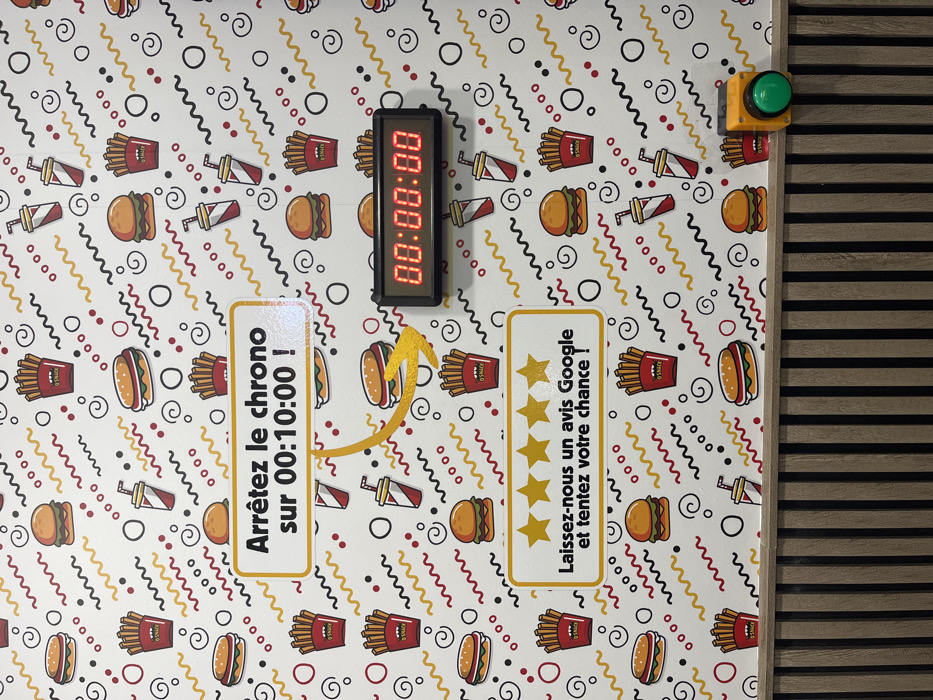 This image captures a section of an interior wall within a fast-food restaurant identified as O'SNACK, located in Strasbourg, France. The wall's upper two-thirds are covered in white wallpaper patterned with cartoon illustrations of hamburgers, french fries (labeled "O'SNACK"), and soda cups, interspersed with red, yellow, and black wavy lines, circles, and dots.
A black digital timer, displaying "00:00:00" in red LED digits, is mounted on the patterned wall. To its upper left, a rectangular white sign with a yellow border instructs in French: "Arrêtez le chrono sur 00:10:00 !" (Stop the timer at 00:10:00!). A yellow, curved arrow points from this sign towards the digital timer. Below this setup, another identical white and yellow sign displays five yellow stars and the French text: "Laissez-nous un avis Google et tentez votre chance !" (Leave us a Google review and try your luck!).
The lower third of the image reveals a wooden slatted wall or counter, composed of alternating light and dark brown vertical planks. A yellow control housing with a prominent circular green push-button is affixed to this wooden structure in the lower right corner. The configuration suggests an interactive promotional game where participants attempt to stop the timer at a specific mark, potentially in conjunction with leaving a Google review. No individuals are present in the scene.