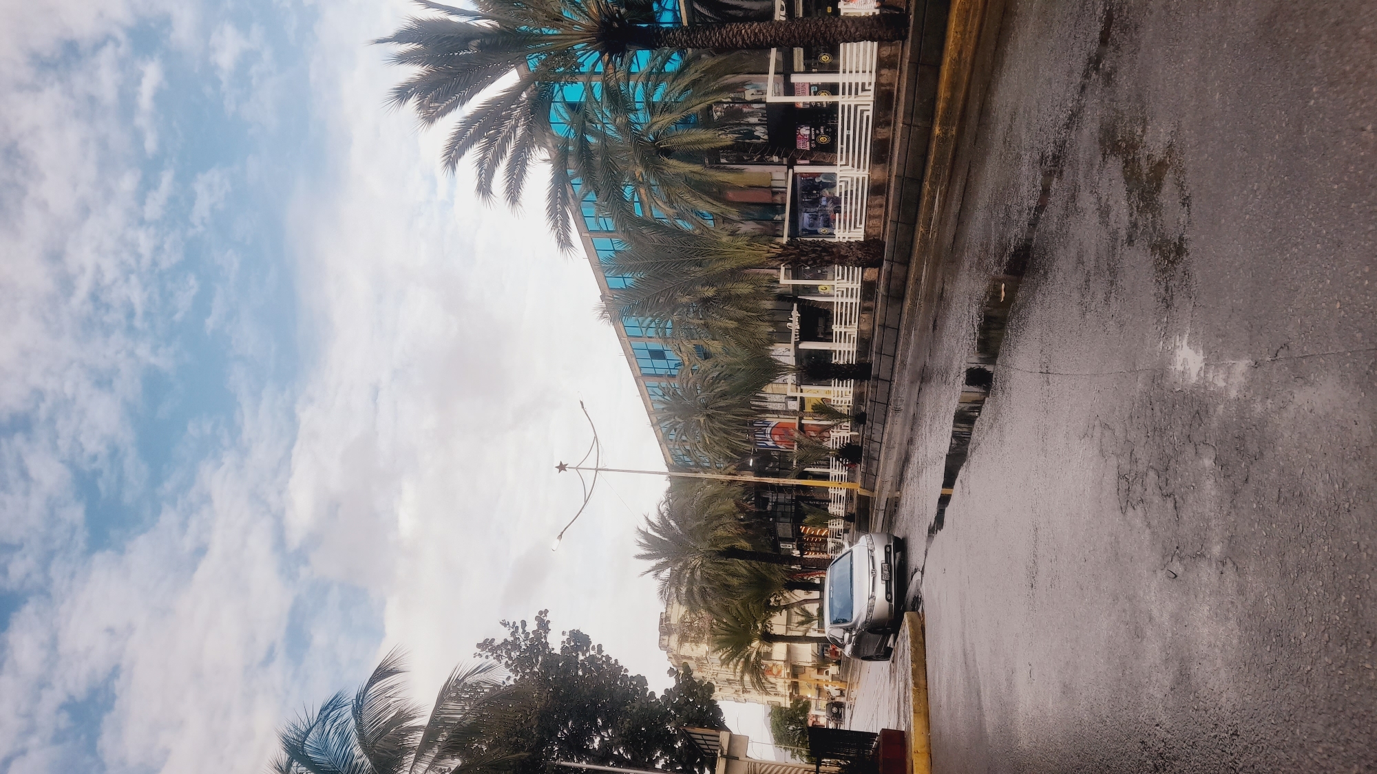 A wet street scene in Juan Griego, Venezuela, with a silver car parked on the left side. The street is wet from rain, with puddles reflecting the cloudy sky. Palm trees line the sidewalk, and a modern building with blue glass windows is visible in the background. The sky is partly cloudy, with a mix of blue sky and white clouds. The overall atmosphere is subdued, with the wet conditions giving the scene a slightly melancholic feel.