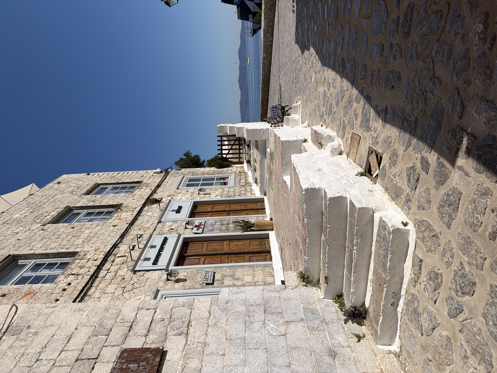This photo shows a street scene in Hydra, Greece. The image captures a stone building with windows and three doors, each with signs above them. Next to the building, a set of white stairs ascends alongside a stone pathway. The scene also captures a glimpse of the sea and a sailboat in the background. The sky is a clear, vibrant blue.