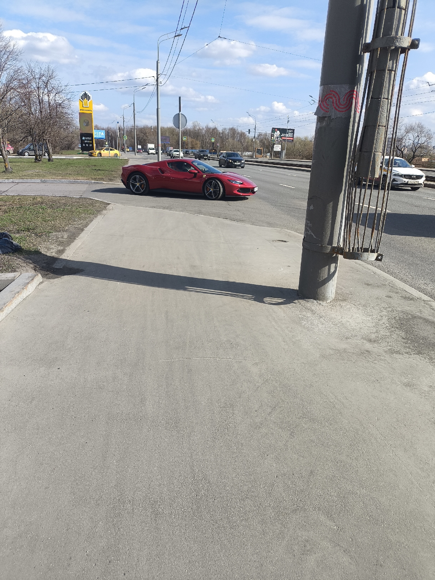The image shows a red Ferrari driving on a road in Moscow, Russia. There is a sidewalk in the foreground and a pole with a metal grate attached to it. Other vehicles are visible on the road. A billboard is visible in the background. The sky is blue with some clouds.