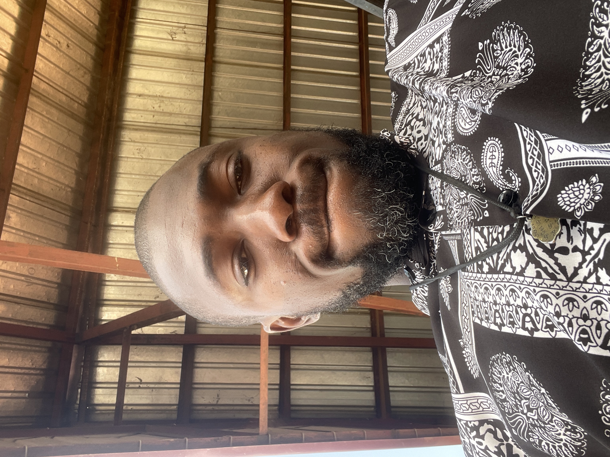A medium close-up portrait shows a Black man with a short haircut and a beard, looking directly at the camera with a slight smile. He is wearing a black short-sleeved shirt with a white paisley pattern and a dark pendant on a string around his neck. The background consists of a corrugated metal roof with exposed wooden beams and framing, suggesting an outdoor or partially enclosed shelter. The lighting is bright, casting shadows and highlighting the textures of the corrugated metal and the man's skin and beard. The scene appears to be in Gura Top, Nigeria, based on the provided context.