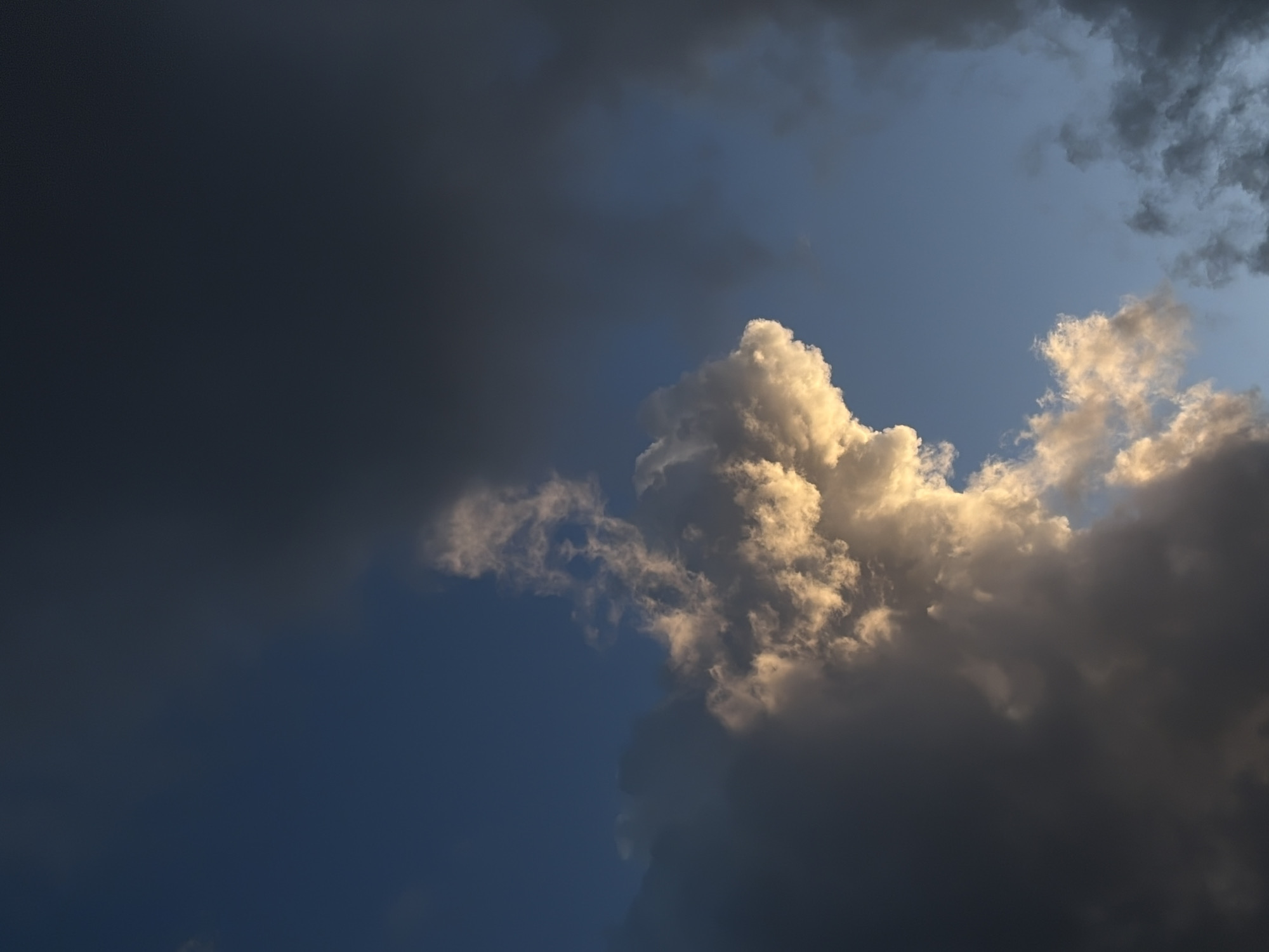 The image presents a sky dominated by contrasting cloud formations. On the left, a dense, dark cloud covers a significant portion of the frame. In the center and right, a large, billowing cumulonimbus cloud is illuminated by sunlight, displaying varying shades of white and grey. The background reveals patches of blue sky, particularly near the center and upper right. The overall impression is that of an approaching storm. The location and any subjects or actions in the photo are not visible, so further detail about the location in Budva, Montenegro, cannot be deduced from the image alone.