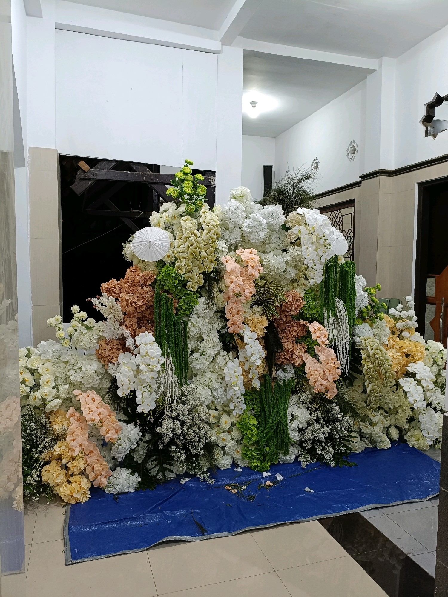 The image shows a large, densely packed floral arrangement with a variety of artificial flowers in shades of white, cream, peach, light yellow, and green. The arrangement is situated indoors, against a white wall with some architectural details, possibly in a hallway or entryway. The flowers are arranged in a cascading manner, filling a significant portion of the frame. Some of the flowers appear to be hydrangeas, orchids, and smaller filler flowers. There are also green, hanging decorative elements that resemble strands of beads or small, hanging plants. A small, white, umbrella-like object is visible within the floral arrangement. The floral display is placed on top of a large, blue tarp that covers the tiled floor. The background includes a doorway and part of a ceiling with lighting fixtures. The location is identified as Maguwoharjo, Indonesia. The scene appears to be set up for an event, possibly a wedding or a celebration. There are no people visible in the image. The lighting is artificial and appears to be coming from overhead fixtures.