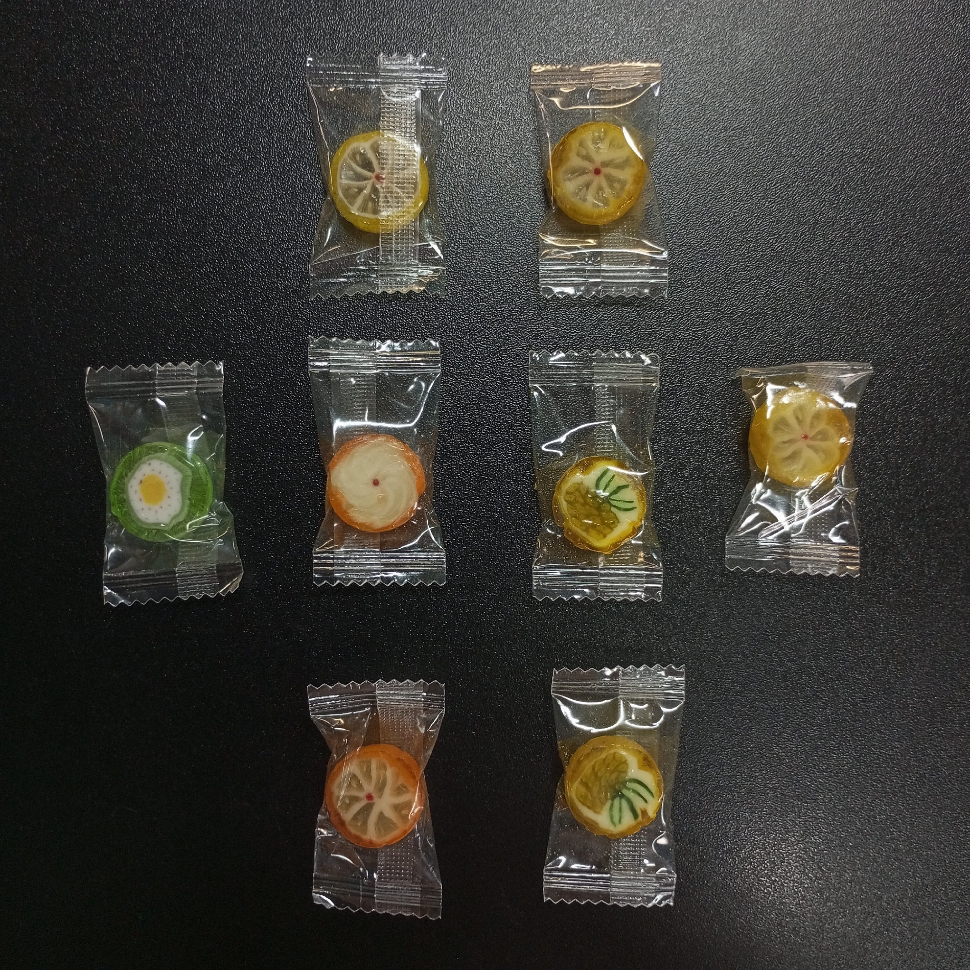 The image shows seven individually wrapped candies arranged on a dark, textured surface. The candies are round and appear to be fruit-flavored, with designs resembling citrus slices. The candies are packaged in clear plastic wrappers with a crimped seal. The background is a dark, flat surface. There is nothing in the image to suggest the location is Pervomaysky, Russia.