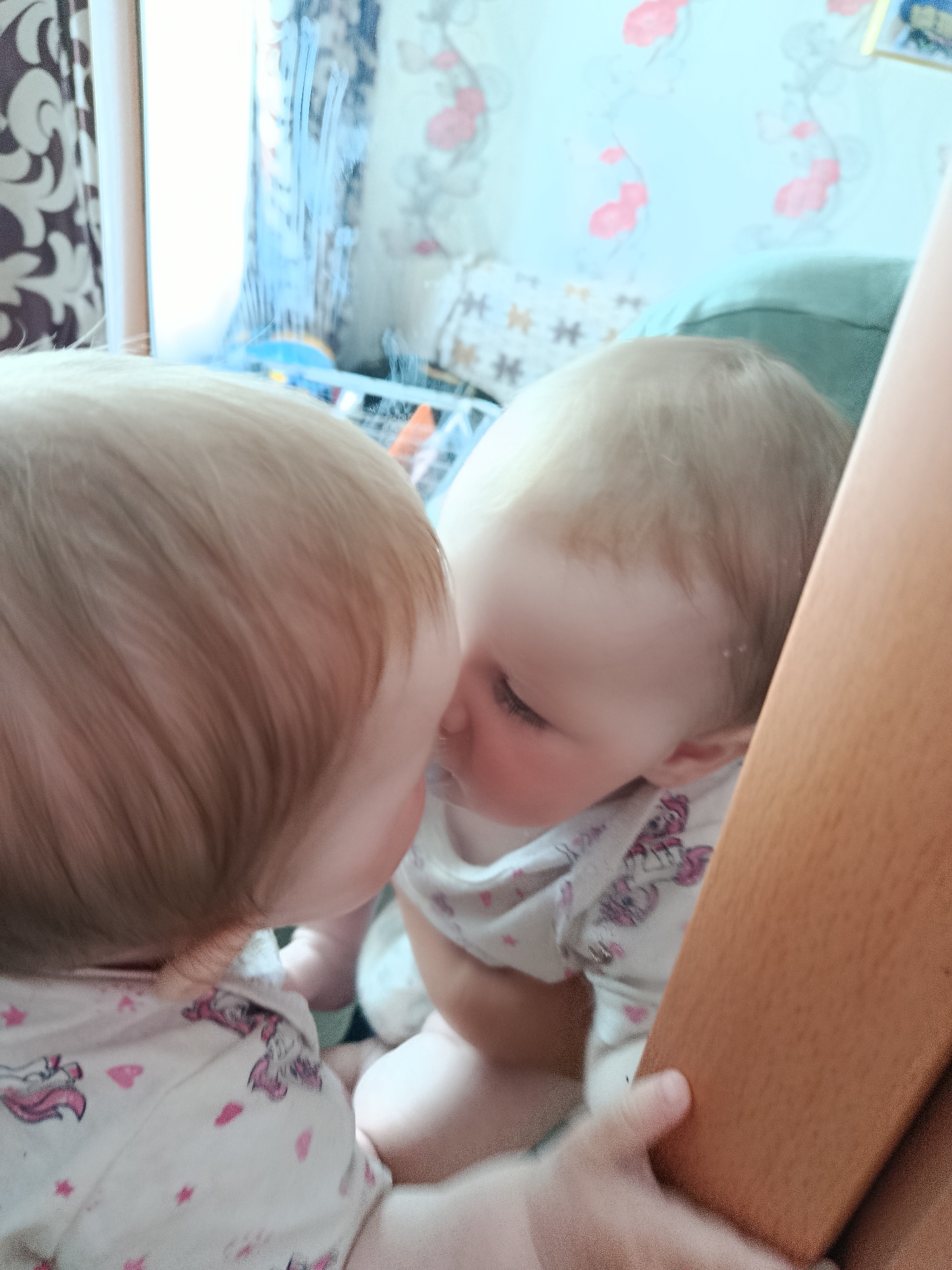 A close-up shot shows a baby interacting with their reflection in a mirror. The baby has light blonde hair and wears a white pajama top patterned with pink unicorns and hearts. The reflection appears to be kissing the baby. In the background, a patterned curtain and wallpaper are visible, alongside the corner of a green pillow and a wooden object. The location is Unknown, Unknown.