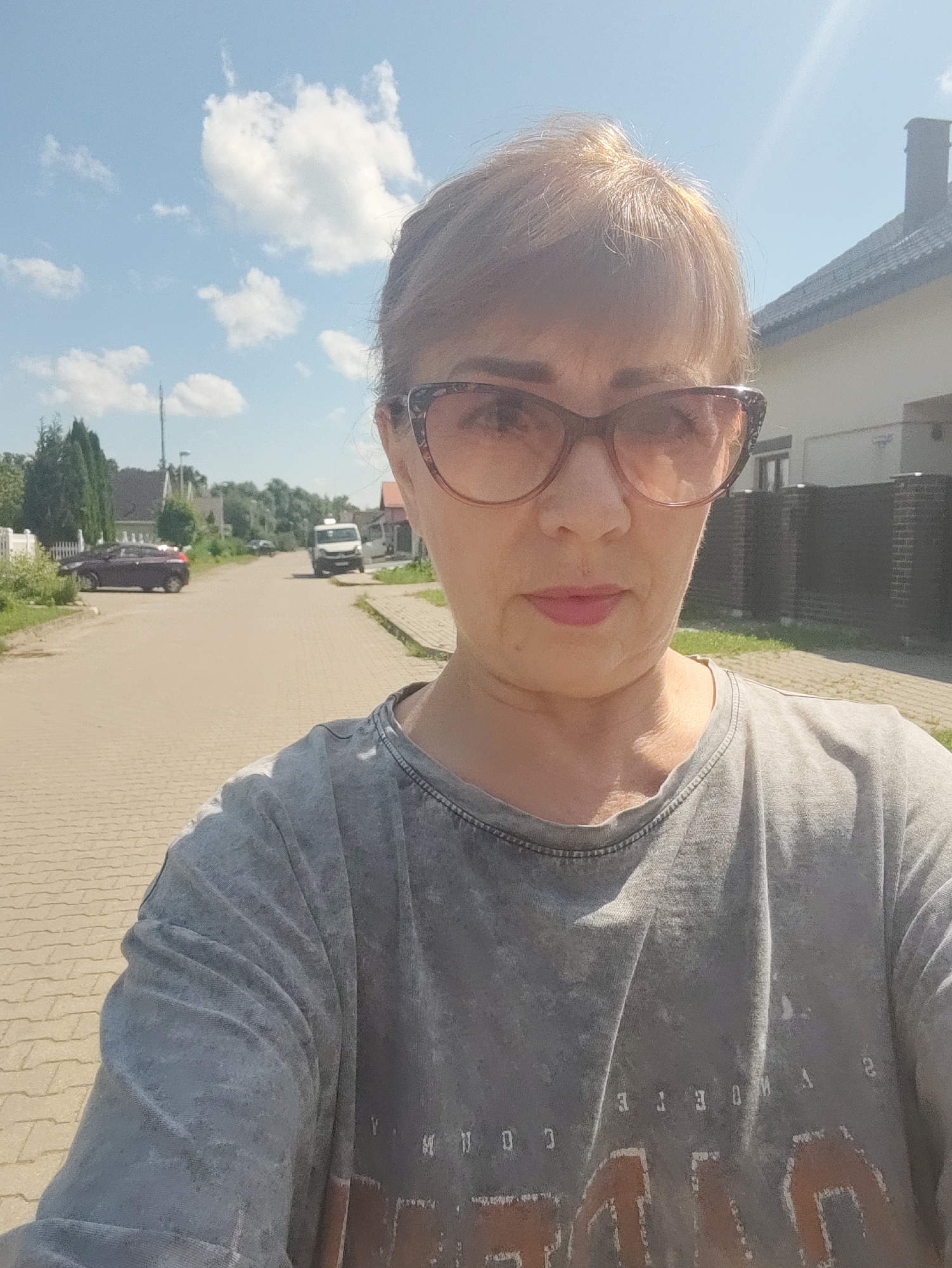 A selfie of a woman wearing sunglasses and a grey t-shirt is taken on a sunny day on a cobblestone street in Gvardeysk, Russia. In the background, there are houses, trees, cars, and a white van driving down the street. The sky is blue with white clouds.