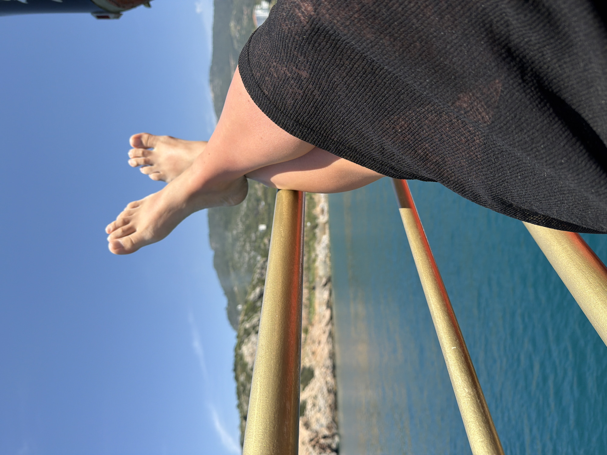 The image captures a serene, low-angle perspective of a person relaxing with their feet elevated, overlooking a scenic coastal landscape. The primary subjects are the bare lower legs and feet of a person, crossed at the ankles, resting upon a golden-colored railing. The person is wearing a sheer, black cover-up or dress which drapes over their thighs, partially obscuring the upper legs. The skin has a healthy, sun-kissed tone.
In the background, a vast, clear blue sky dominates the upper portion of the scene, indicating bright, sunny weather. Below the railing, calm, blue-green sea water stretches out, reflecting the sky. In the mid-distance, a rugged, rocky coastline adorned with some greenery is visible across the water, with mountainous terrain further inland. The perspective suggests the person is on a boat, a balcony, or a high vantage point, enjoying the view.
The activity depicted is one of leisure and relaxation. The bright lighting points to a daytime scene. The combination of clear blue water, a rocky coastline, and mountains is consistent with the Mediterranean environment of Alanya, Turkey. No text is visible in the media file.