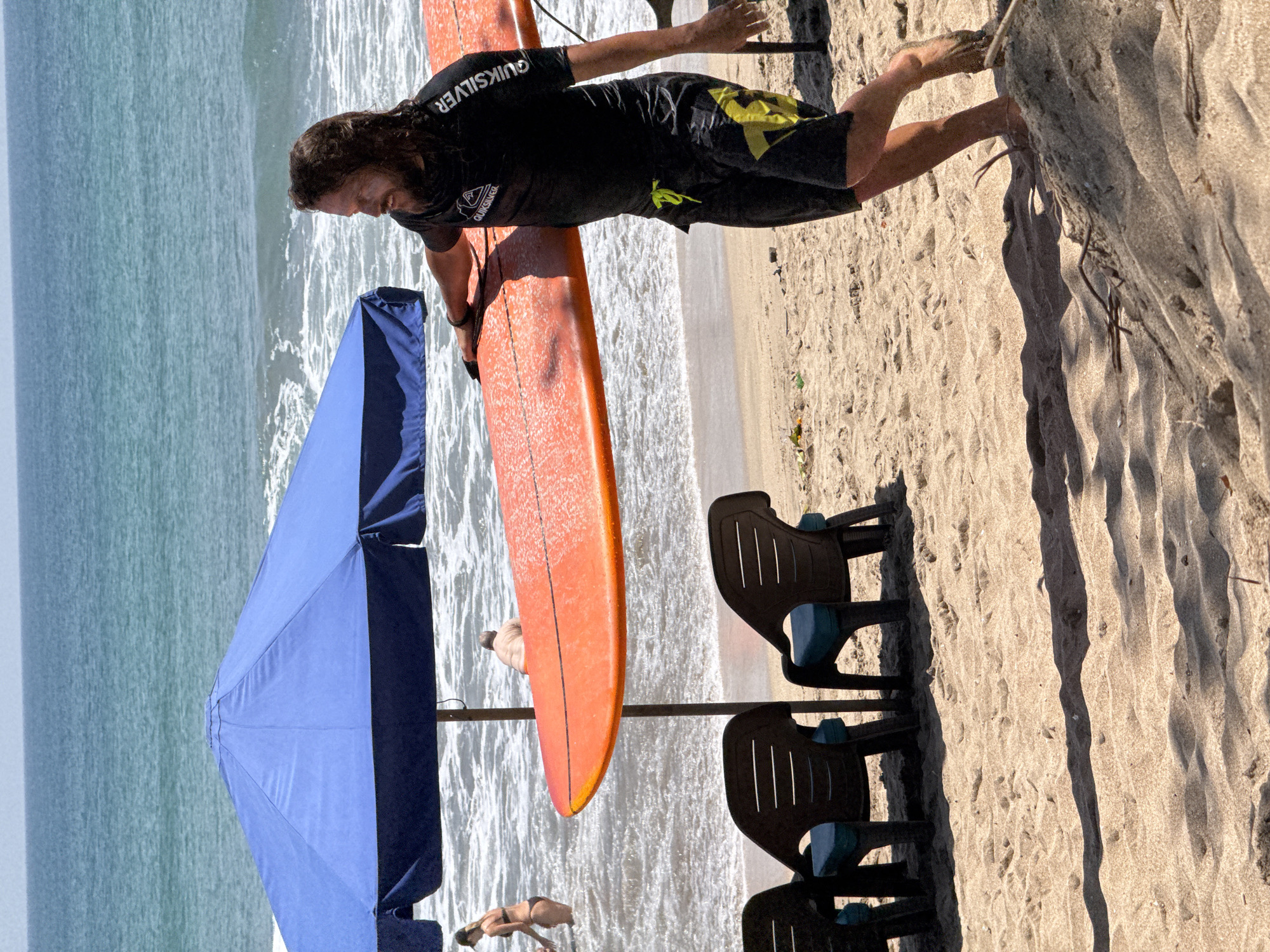 In Kuta, Indonesia, on a sandy beach, a man with long, dark, wavy hair and a beard walks towards the right, carrying an orange surfboard. He is wearing a black Quiksilver rashguard and black swim trunks with yellow accents. His left hand rests on the surfboard, and his right arm is extended slightly behind him. In the background, the ocean waves crash onto the shore under a clear sky. A large blue umbrella shades a portion of the beach, and two dark plastic chairs with blue cushions are visible in the foreground. To the left of the umbrella, a woman in a black bikini walks in the shallow water, and further back, a person with their back to the camera stands in the ocean. The sand is textured with footprints and scattered debris.