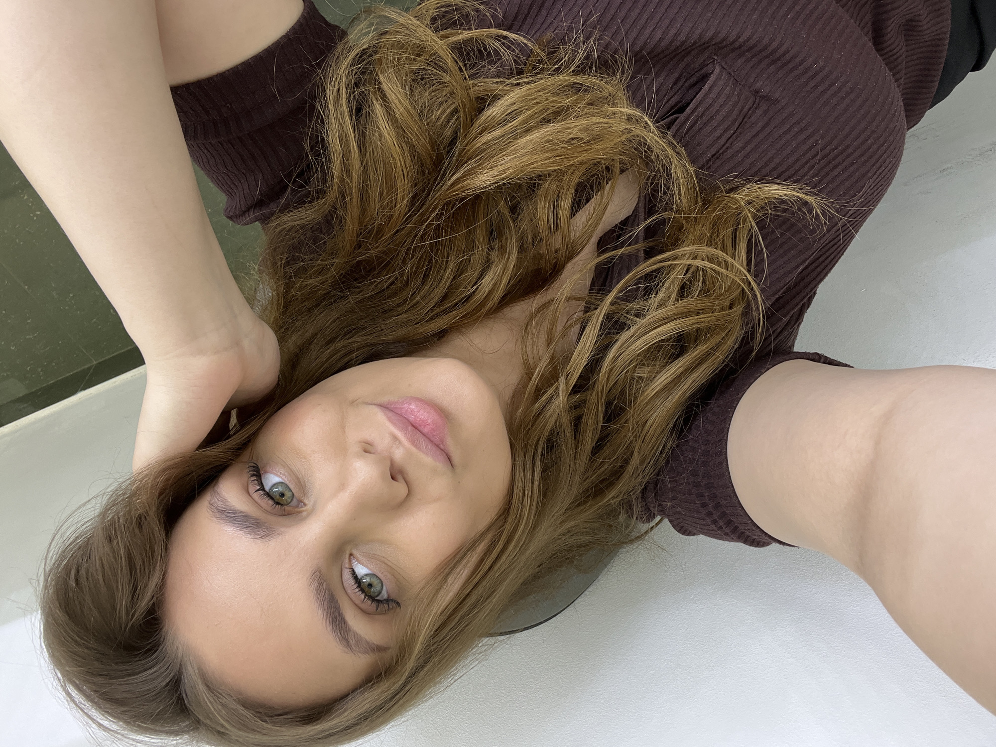 A woman with fair skin and light brown hair is lying down. She is wearing a brown ribbed top. Her right hand is touching her hair. The background is a white surface and part of a green surface. The location is unknown and there is no indication the image was taken in Chaikovsky, Russia.
