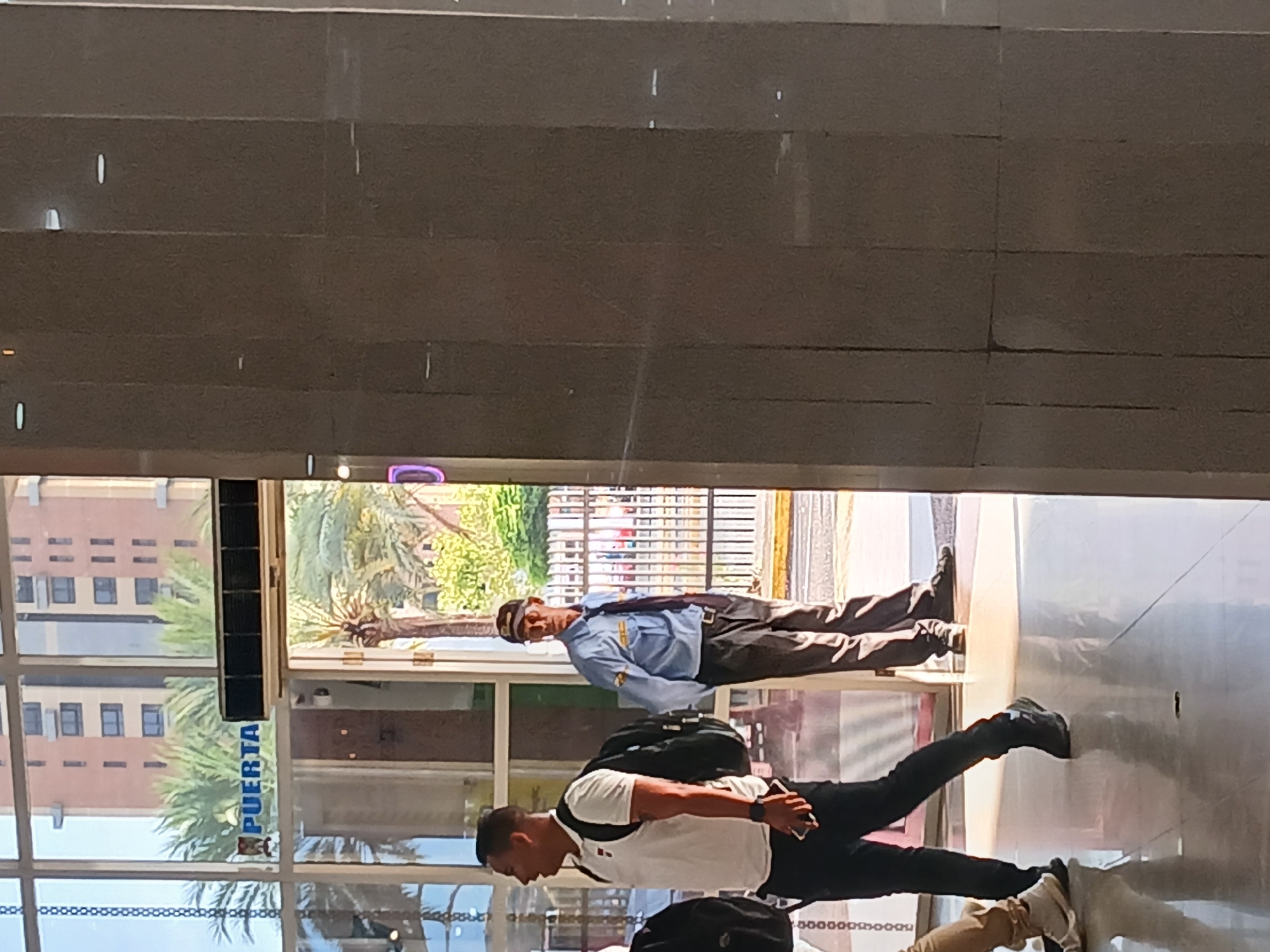 The image shows the interior of a building in Ciudad Guayana, Venezuela, likely a shopping mall or commercial center, due to the presence of exterior glass doors. A security guard wearing a light blue shirt, dark pants, and a cap is standing in the doorway. Another person, wearing a white t-shirt and black pants, with a black backpack, is walking out. The word "PUERTA" can be seen on the glass doors to the left of the frame. Across the street, a tall building is visible. The environment is sunlit and the scene seems busy with people passing.