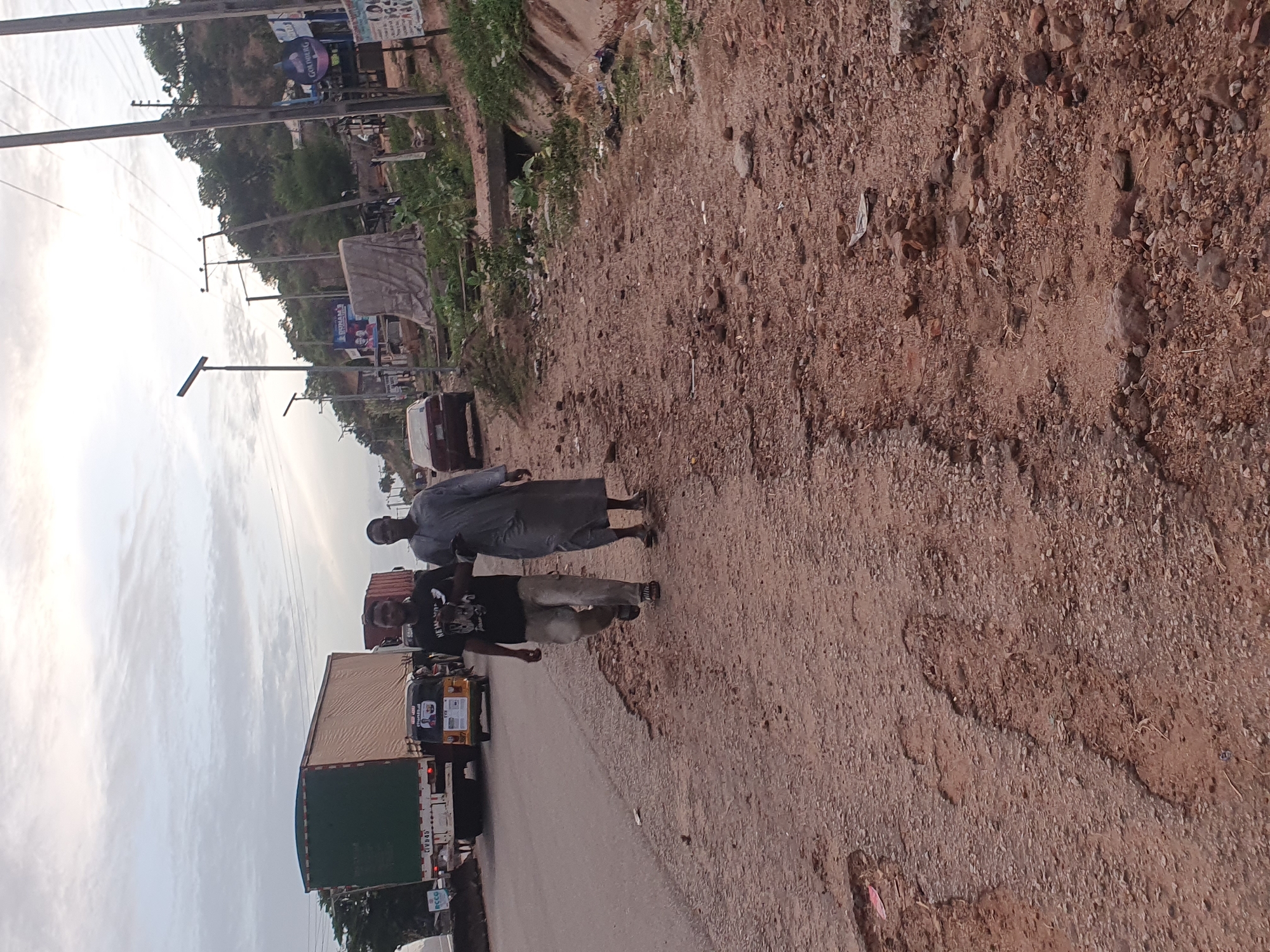 The image shows a road in Lokoja, Nigeria, with two people walking along the side. A truck and a tricycle are on the road. The roadside is unpaved and has loose dirt and rocks. Utility poles and buildings are visible in the background. The sky is overcast.