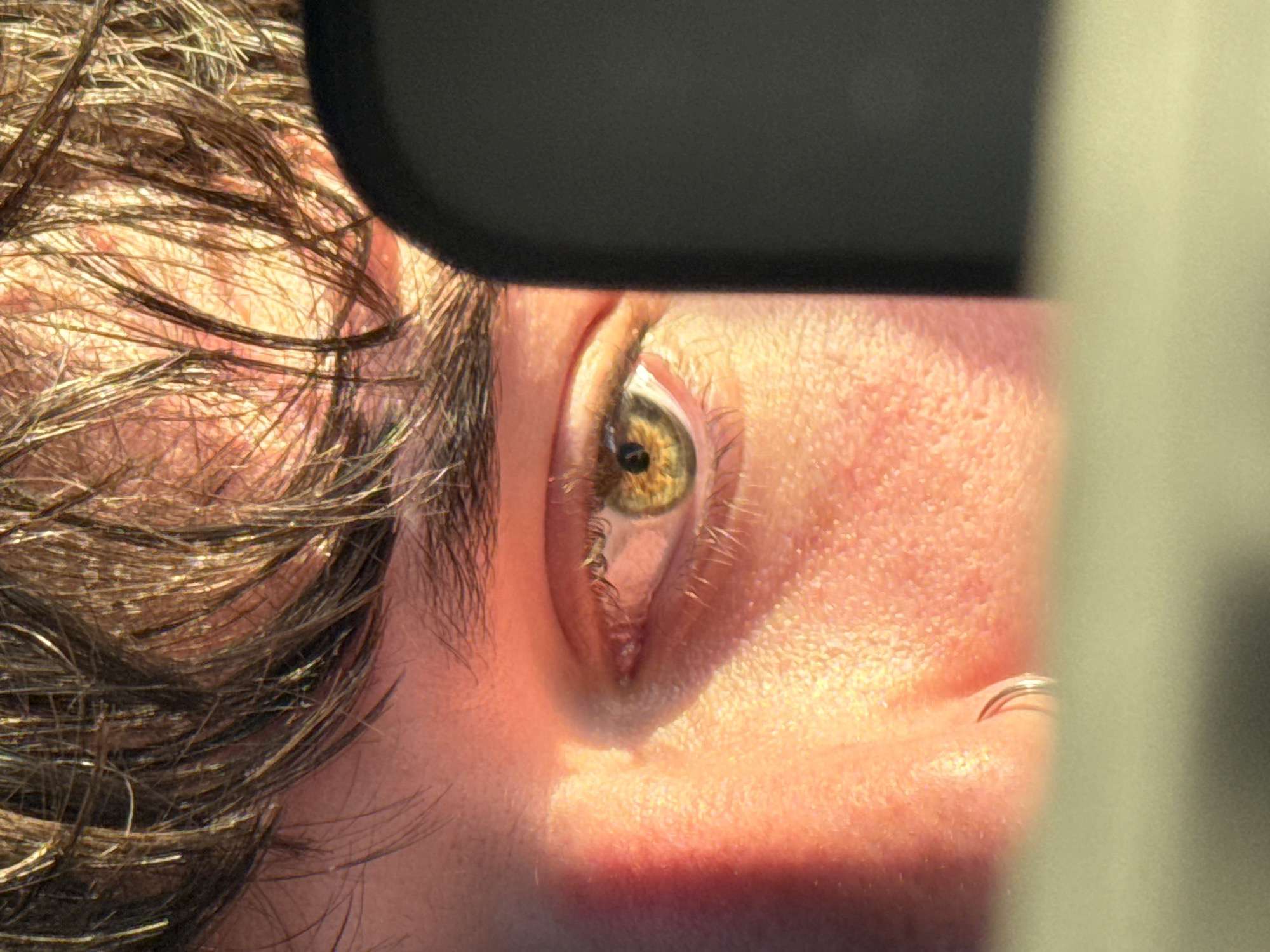 The image presents a close-up of a person's face, focusing on the right eye. The eye is hazel in color, with visible eyelashes and surrounding skin exhibiting pores and texture. The person has brown hair, some of which is visible near the temple. A portion of what appears to be a black sunglasses frame is present at the top of the image. A silver nose ring is visible. The environment or location is undetermined based on the visual information provided.