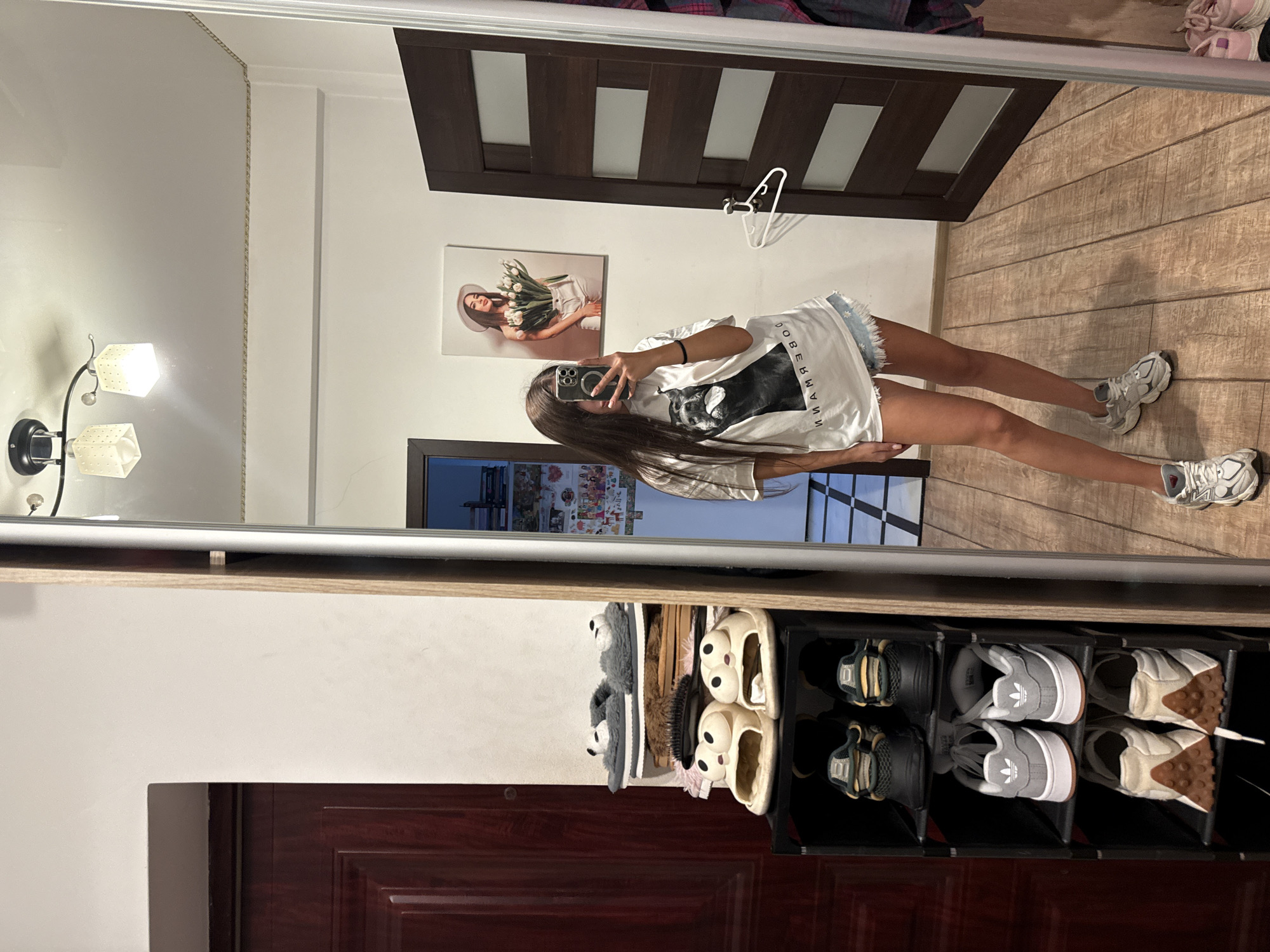 The image depicts an indoor scene, likely a residential hallway or entryway, based on the presence of a shoe rack, doors, and a mirror. The overall lighting suggests it is daytime, possibly indoors with artificial lighting.
In the foreground, a young woman is taking a mirror selfie. She has long, dark hair and is wearing a white t-shirt with a black dog graphic and the word "DOBERMANN" printed on it, paired with light blue denim shorts. She is also wearing athletic shoes.
To the left of the woman, a black multi-tiered shoe rack is visible, filled with various footwear including slippers and athletic shoes. Behind the shoe rack, a dark wooden door is partially visible.
To the right of the woman and slightly behind her is a dark wooden door with horizontal glass panels. A white hanger is attached to the door.
In the background, a large mirror reflects the scene, showing the woman and the shoe rack. On the wall above and behind the woman, a framed picture of a woman holding flowers is displayed. Further in the background, more shelving and a wall decorated with various items, possibly posters or photos, can be seen through a doorway. The floor is made of wooden planks.
The user's request to include the "location in the city of Chișinău, Moldova" cannot be fulfilled as there are no visual cues in the image that would allow for such a determination.