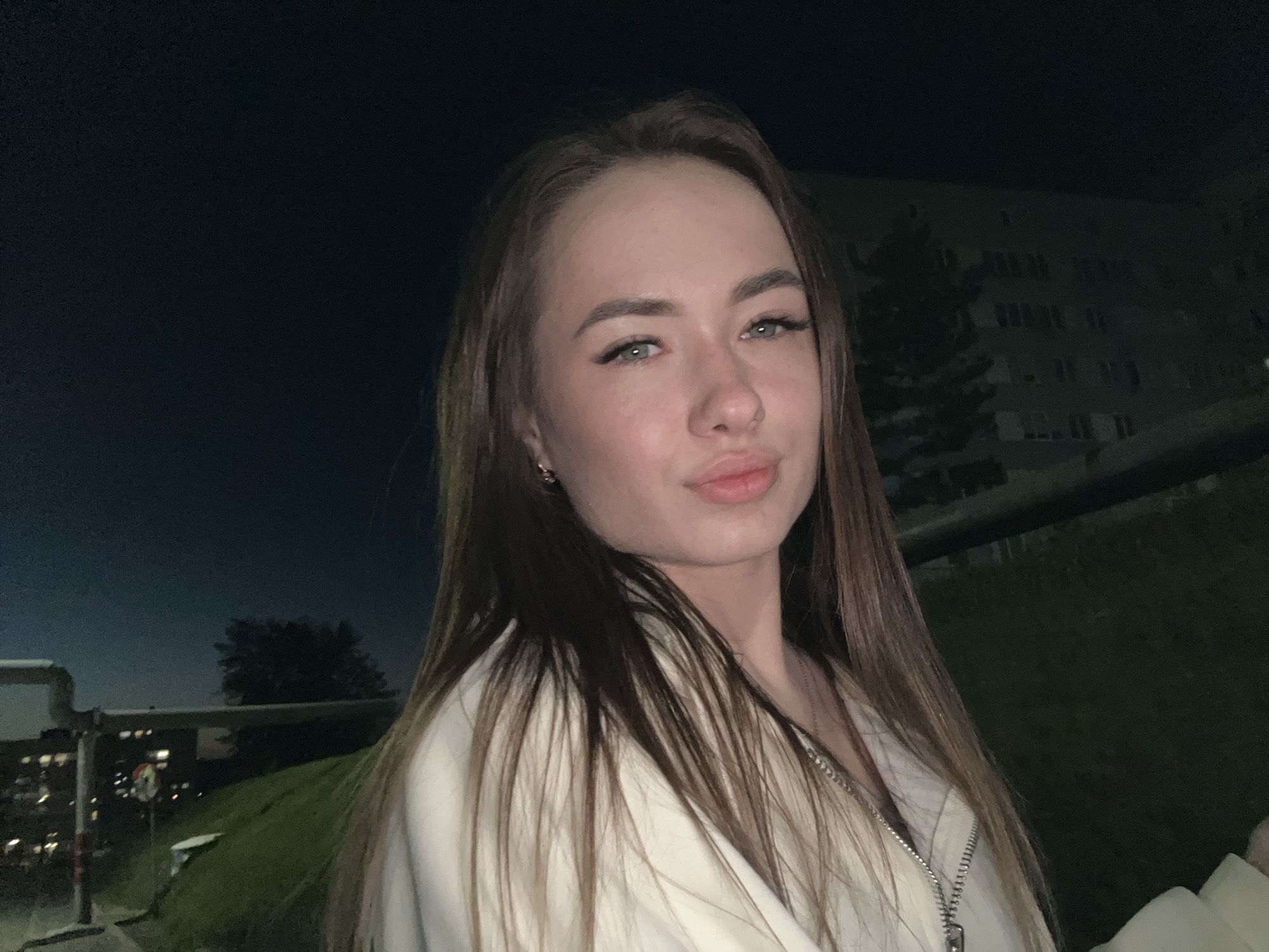 A close-up, eye-level outdoor shot features a young woman centered in the frame against a dark, nighttime backdrop. She has long, straight brown hair parted in the middle, extending over her shoulders. Her face is well-lit, highlighting her light-colored eyes, defined eyebrows, and lips posed in a slight pout. She wears false eyelashes. In her left ear, a small gold-colored hoop earring is visible.
She is dressed in a light-colored (possibly white or off-white) jacket or hoodie with a prominent silver zipper running down the front. A thin silver chain necklace is also visible.
The background is dark, indicating night. On the left side, faint lights of what appear to be distant buildings or vehicles can be seen, along with some industrial-looking pipes or structures. A grassy slope rises towards the center-right. Further back on the right, a large, dark building with numerous faint windows is visible, partially obscured by a dark tree. The overall atmosphere is dim, with the woman being the primary source of light and focus. No discernible text is visible in the image.