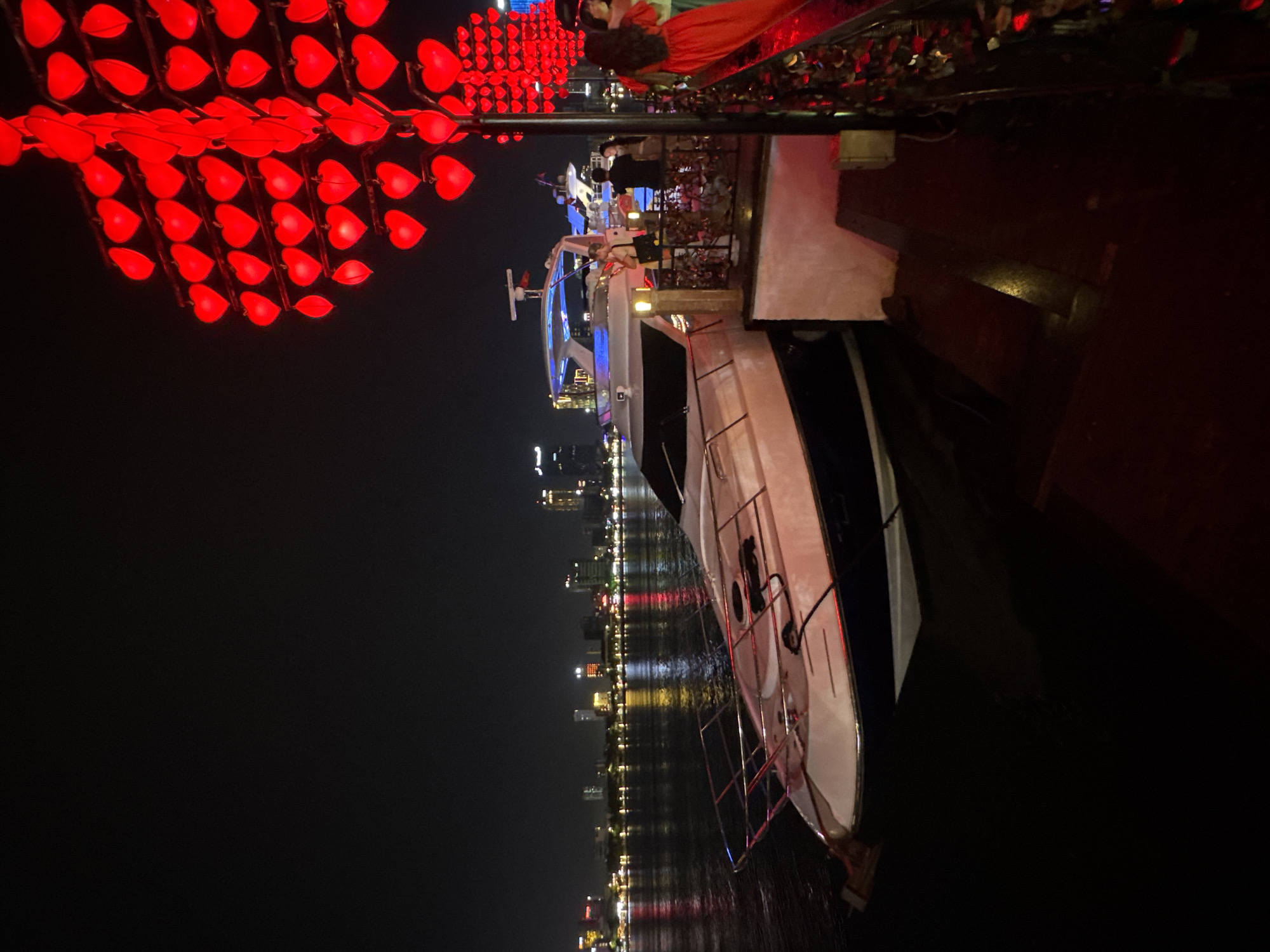 The image shows a nighttime scene in Đà Nẵng, Vietnam. A large yacht is docked alongside a pier or embankment. People can be seen on the upper deck of the yacht. A display of red heart-shaped lights is positioned above the pier, adjacent to the yacht. Across the water, the city skyline is visible with lighted buildings. The water reflects the city lights.