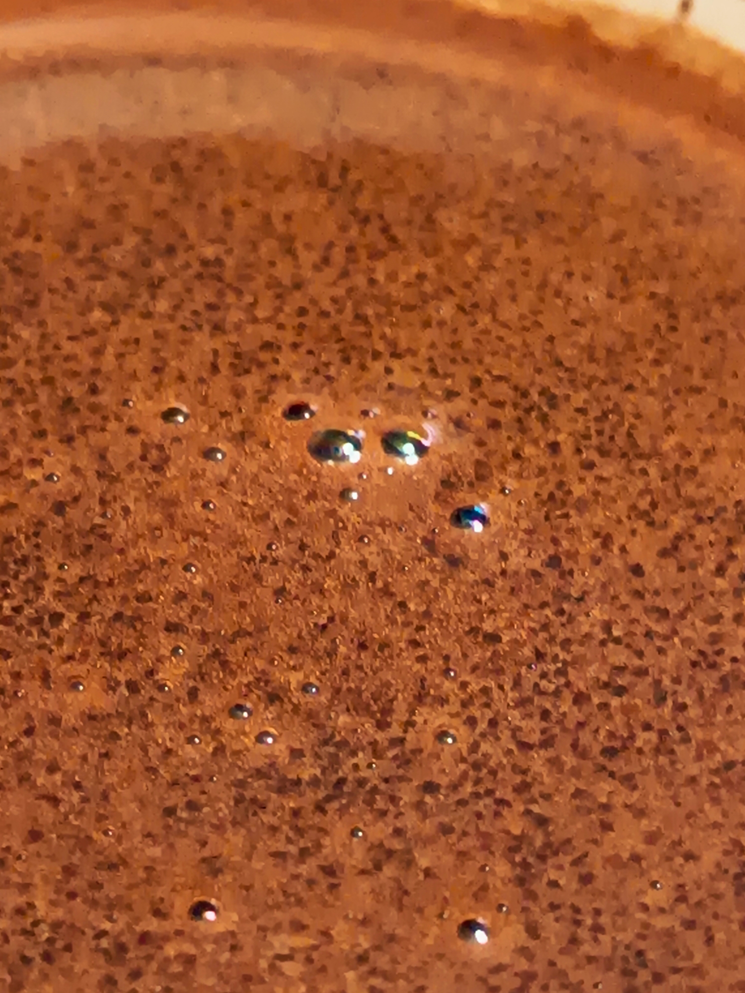 The image is a close-up, overhead view of a dark, bubbling liquid, likely coffee or a similar beverage. The surface is textured with small, dark particles suspended in the liquid, and numerous small bubbles are visible, some reflecting light with iridescent hues. A lighter, smoother area is visible at the top of the frame, possibly the rim of a container. The image does not depict any identifiable landmarks, street scenes, or activities within the city of Izmail, Ukraine.